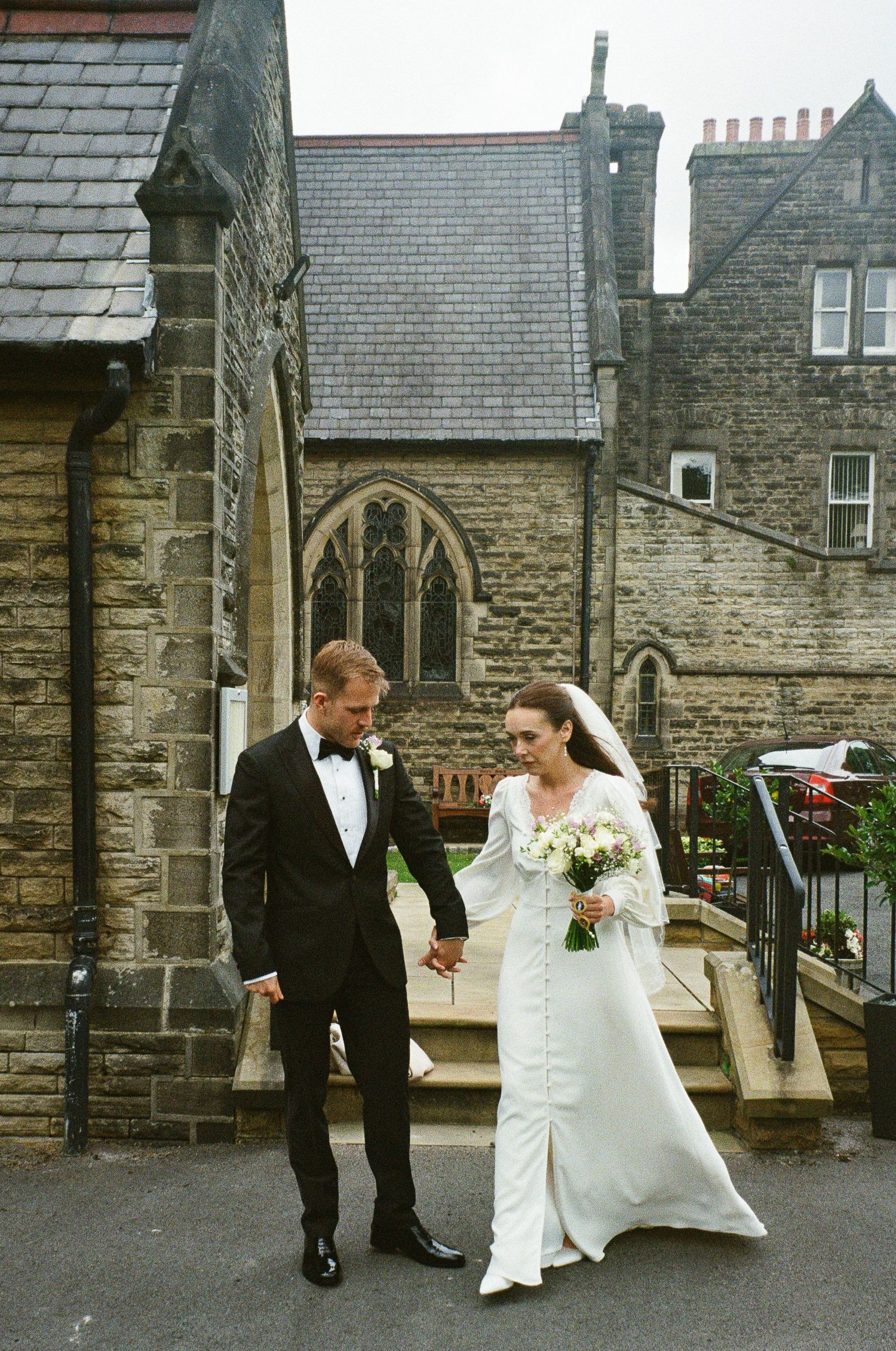 Bride and groom holding hands outside a historic stone church, bride in white gown with bouquet, groom in black tuxedo.
