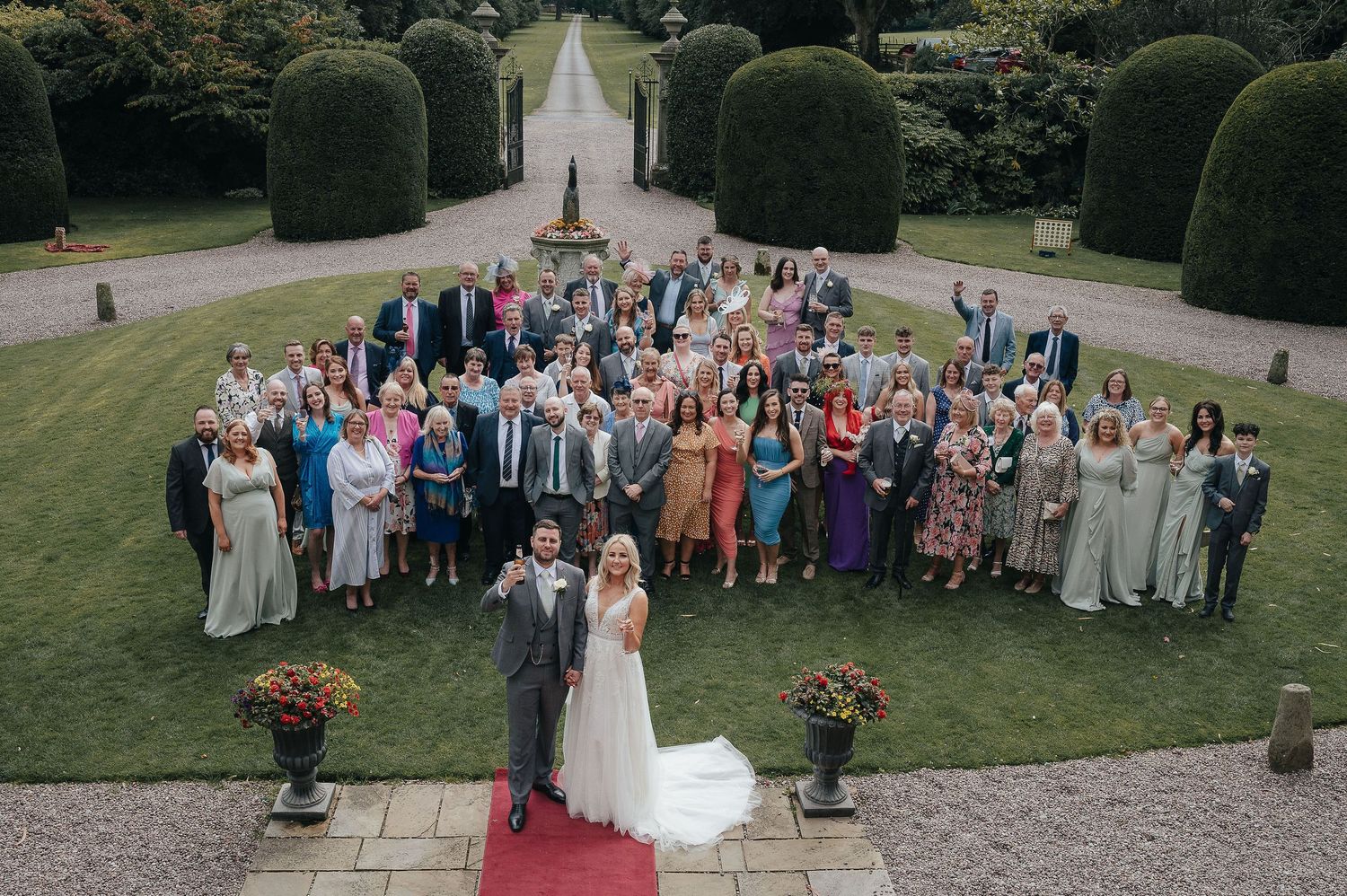 Aerial view of wedding party and guests gathered for group photo on manicured garden grounds.