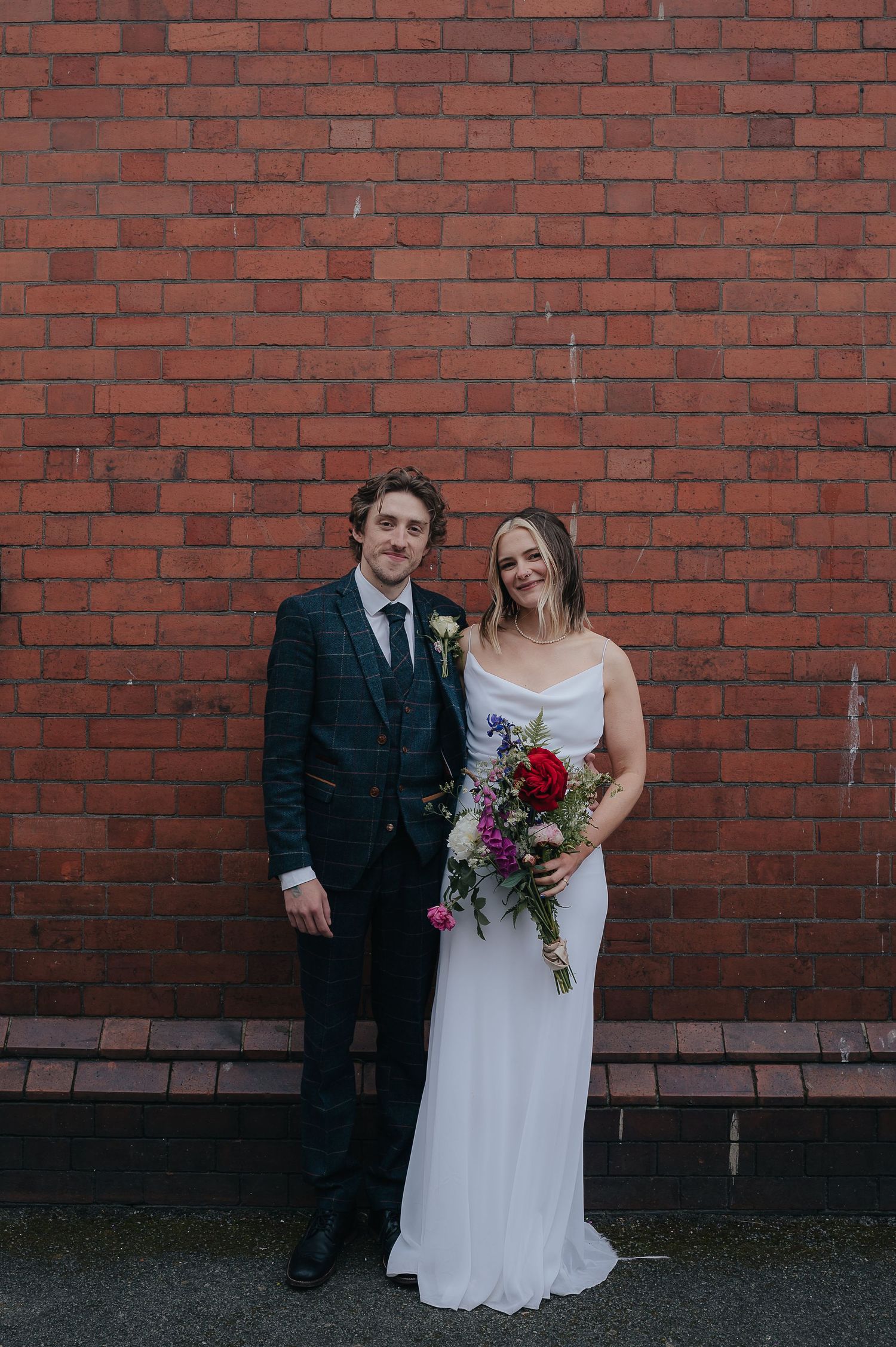 A wedding couple poses against a red brick wall with the bride holding a vibrant bouquet.