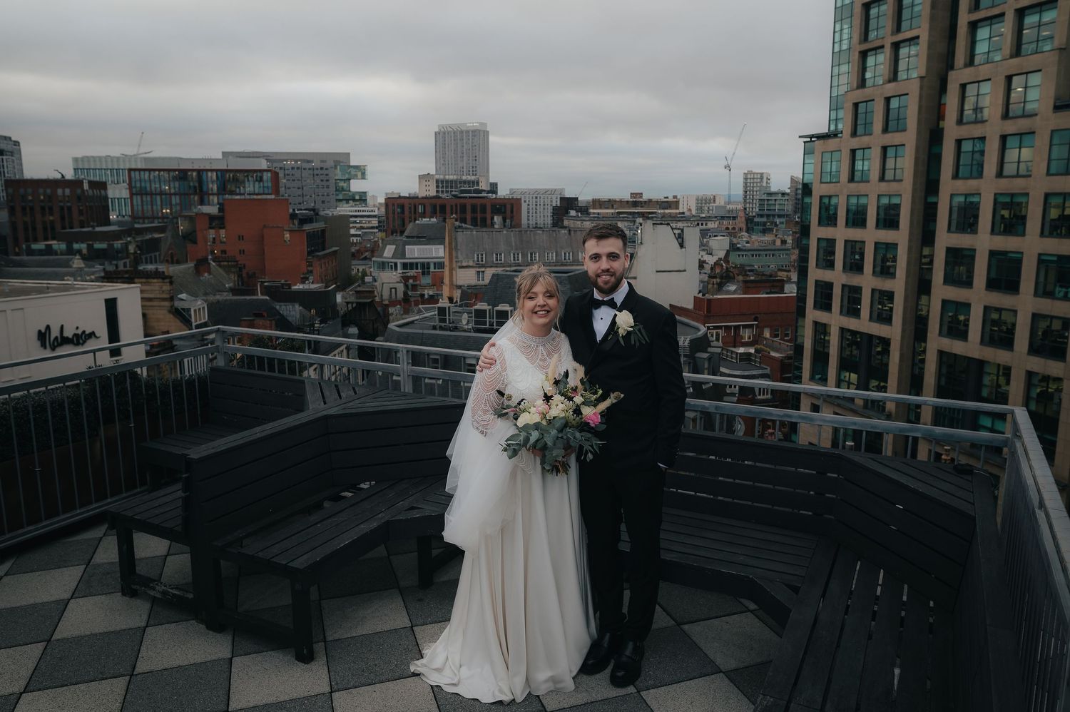 A wedding couple stands on a rooftop deck overlooking an urban cityscape on a cloudy winter day.