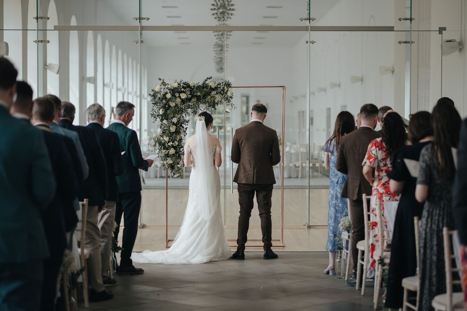 Wedding ceremony in a minimalist white hall with guests seated on either side.