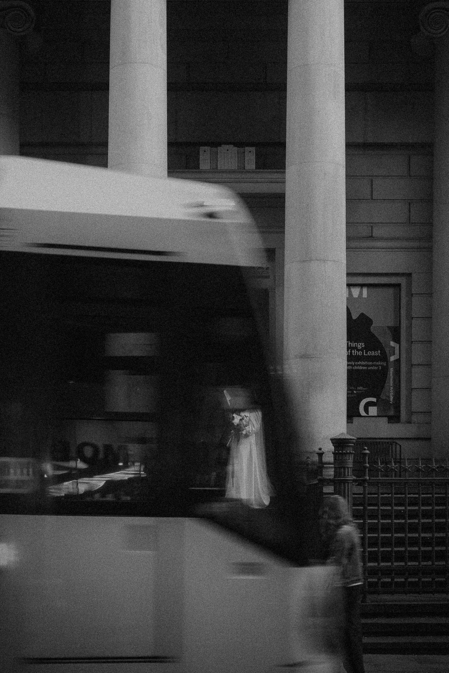 Black and white photo of a blurred tram passing a grand columned building with a figure in white dress visible behind.