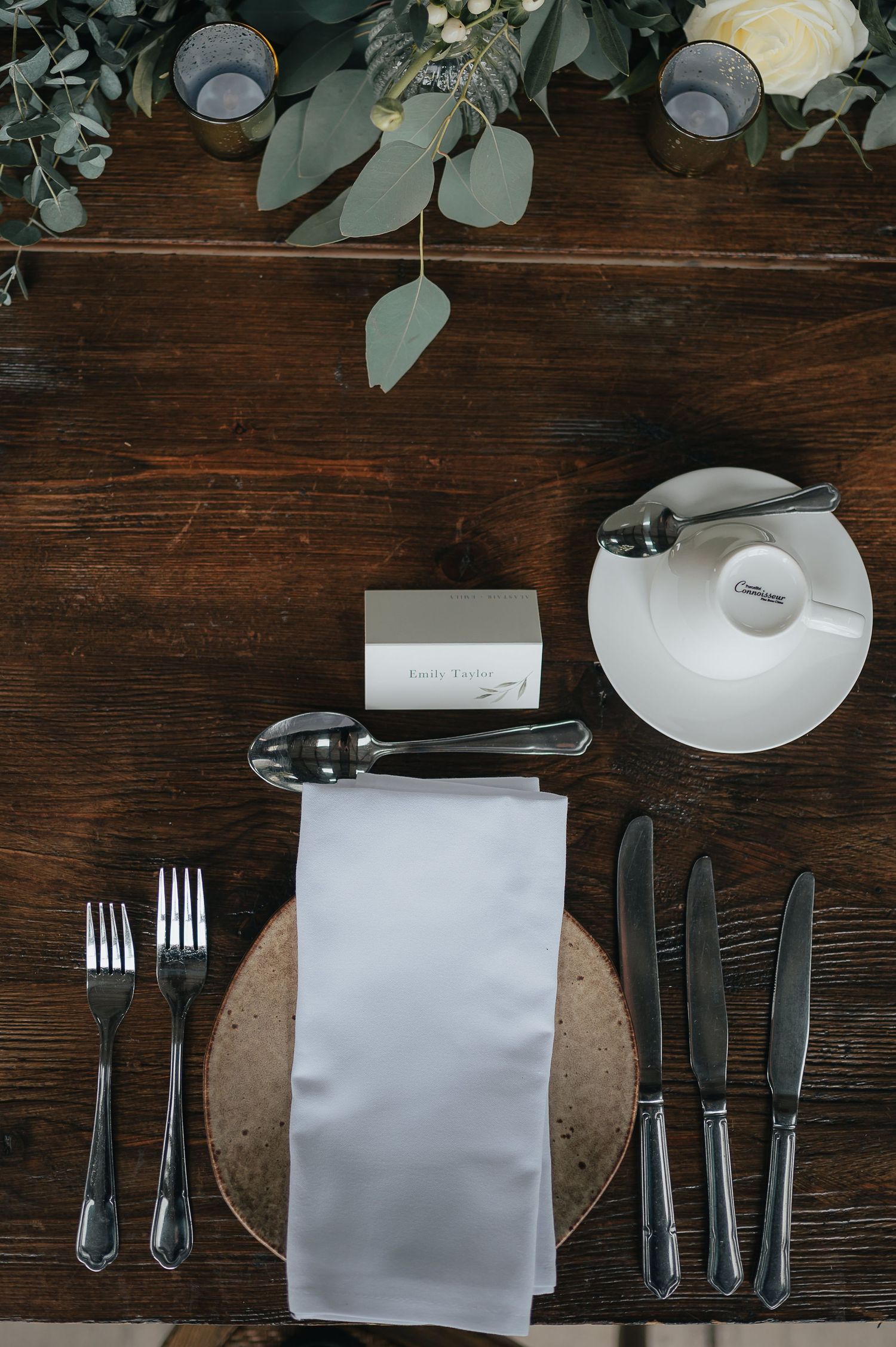 Overhead view of formal place setting with silverware and greenery on dark wooden table.