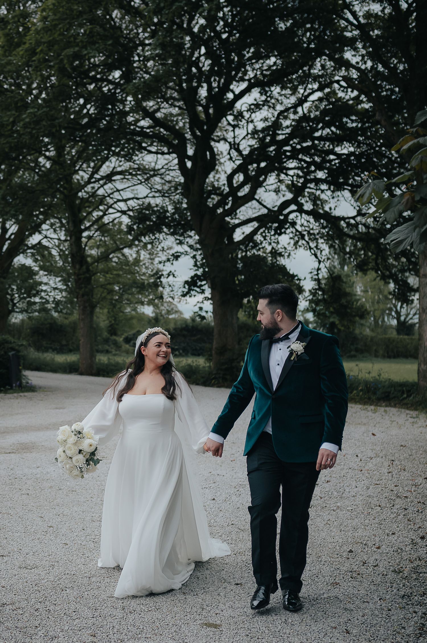 A couple walks hand in hand on a gravel path during their wedding day surrounded by trees.