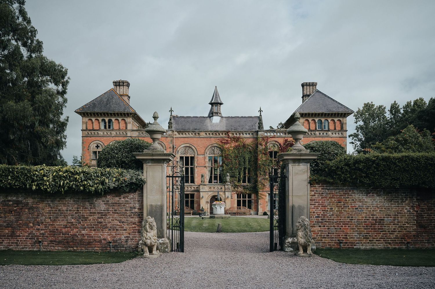 Grand brick mansion with ornate entrance gates and stone pillars set against a cloudy sky.