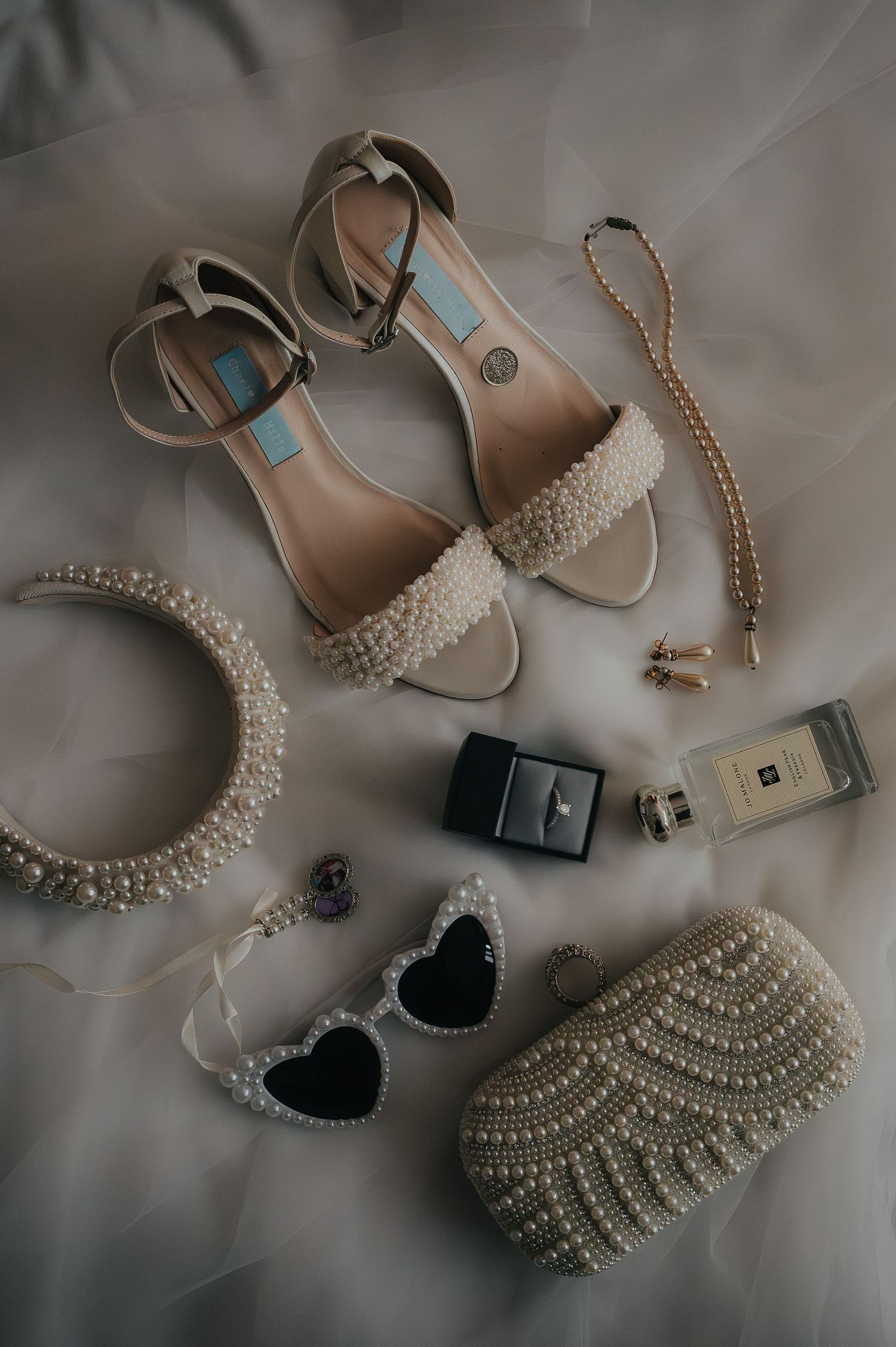 Elegant bridal accessories including pearl-adorned sandals, heart sunglasses, perfume bottles, and beaded jewelry on a neutral surface.