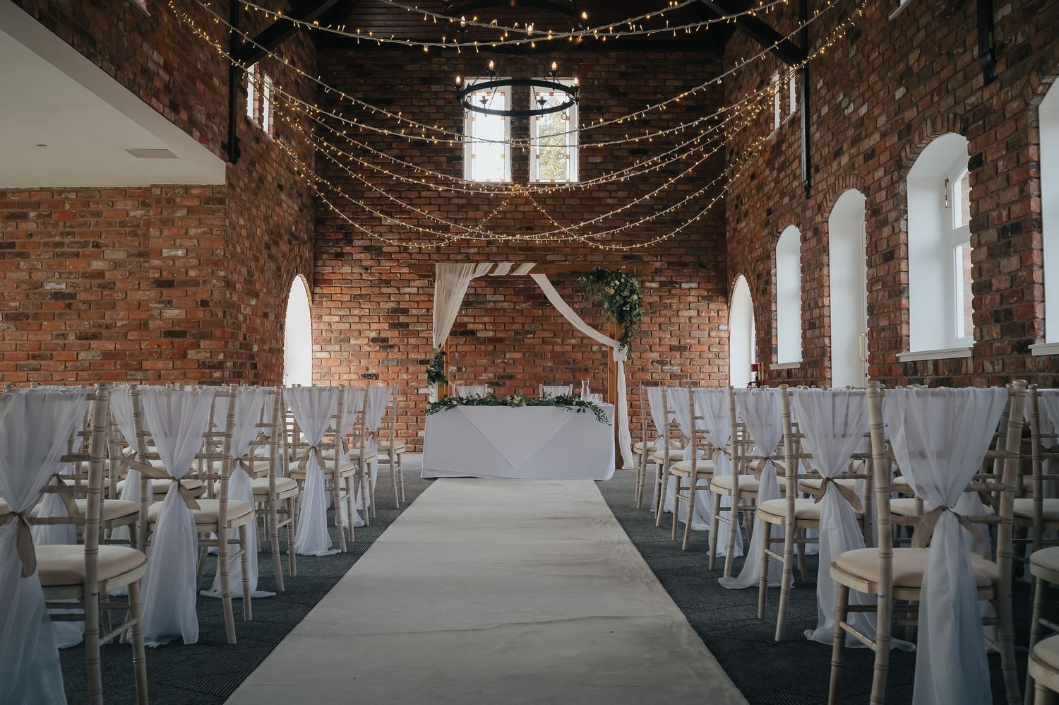 Rustic brick wedding venue with white chairs and a wooden arch decorated with fairy lights and white fabric.