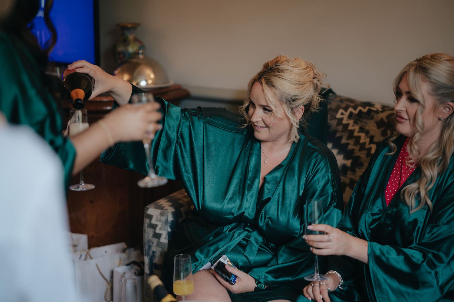 Wedding party members in emerald green robes share drinks and laughter during pre-ceremony preparations.