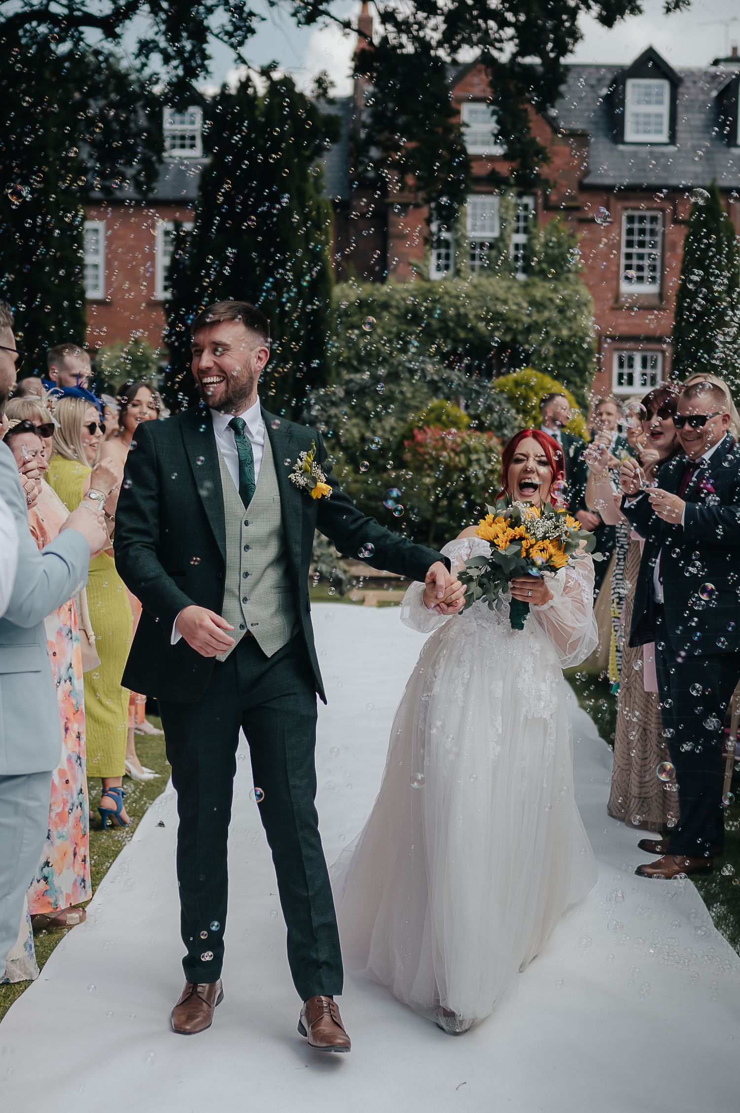 Newlyweds walking down white aisle runner during recessional while guests throw confetti.