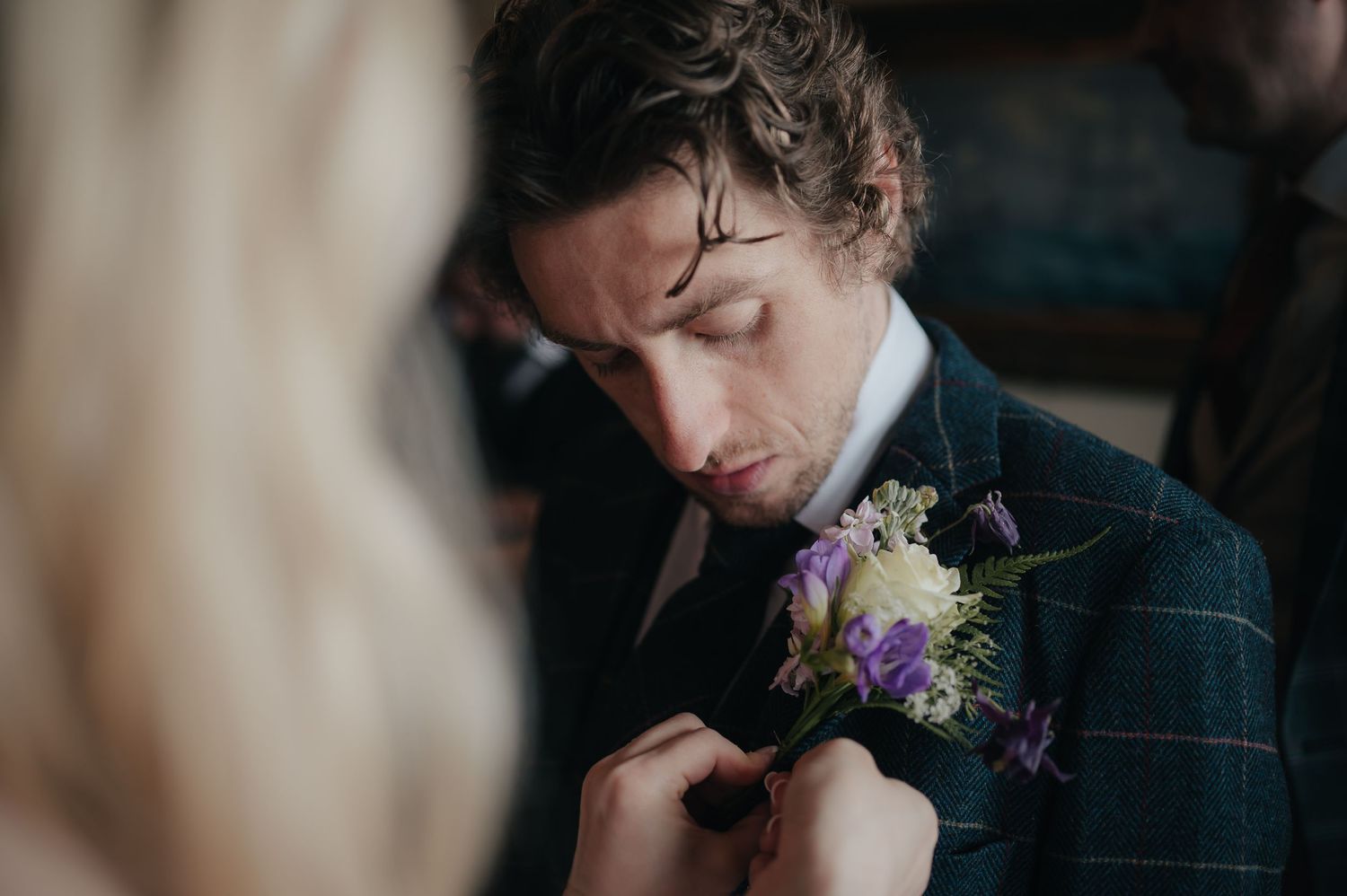 A person in a dark suit adjusting a floral boutonniere with purple flowers.