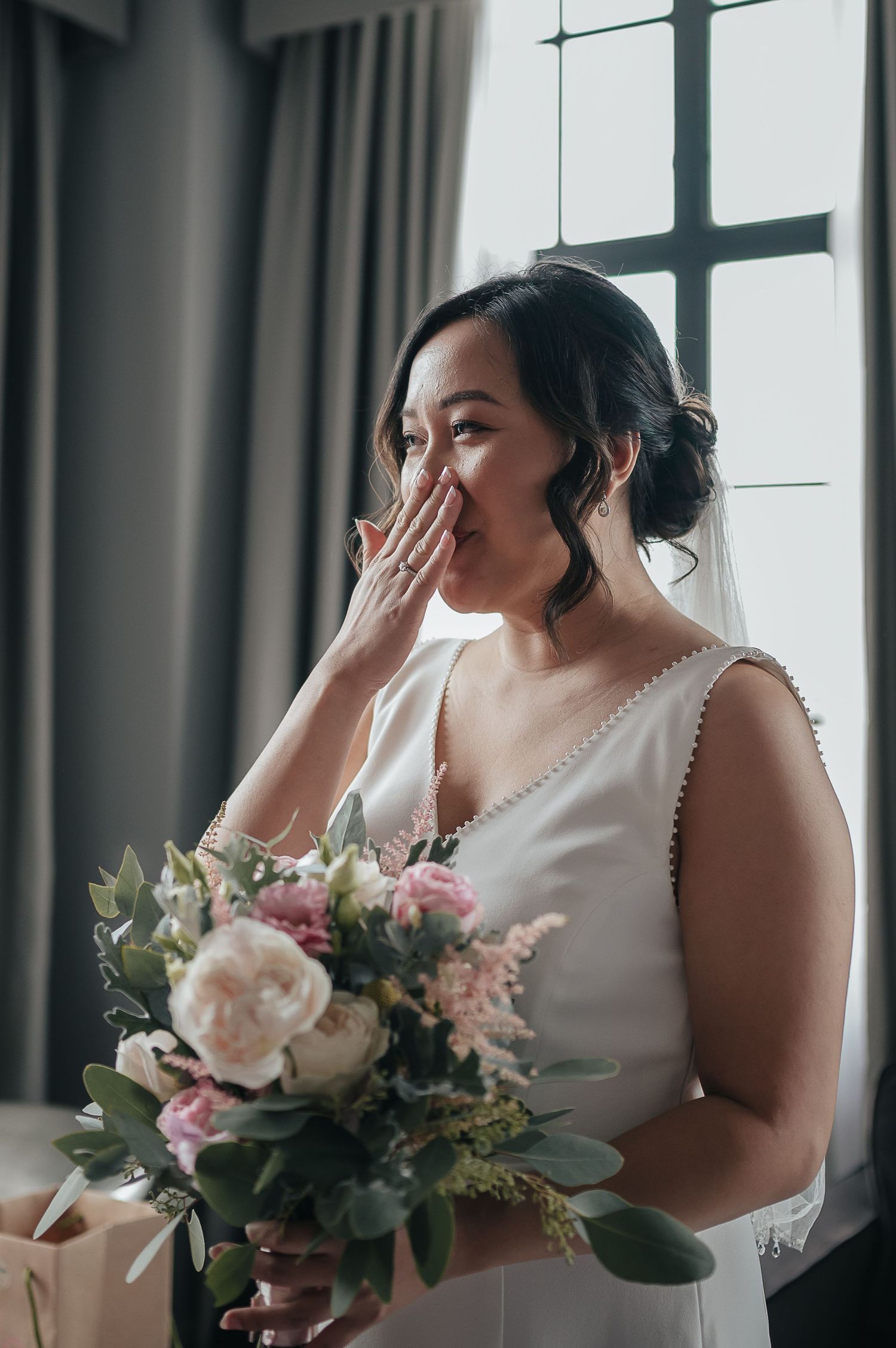 Emotional bride in white dress holding bouquet by window during wedding preparation.