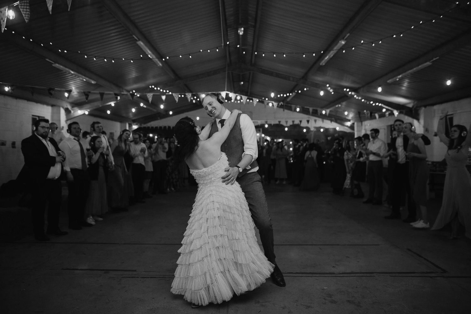 A couple shares their first dance at a wedding reception beneath twinkling string lights in a rustic venue.
