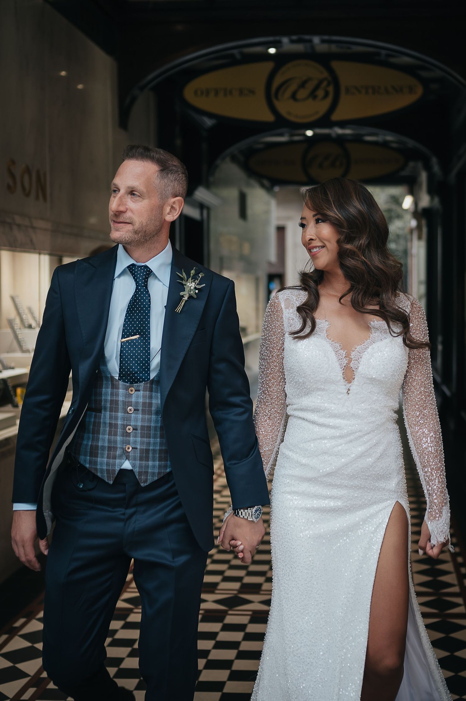 Couple in elegant wedding attire walk under vintage storefront entrance.