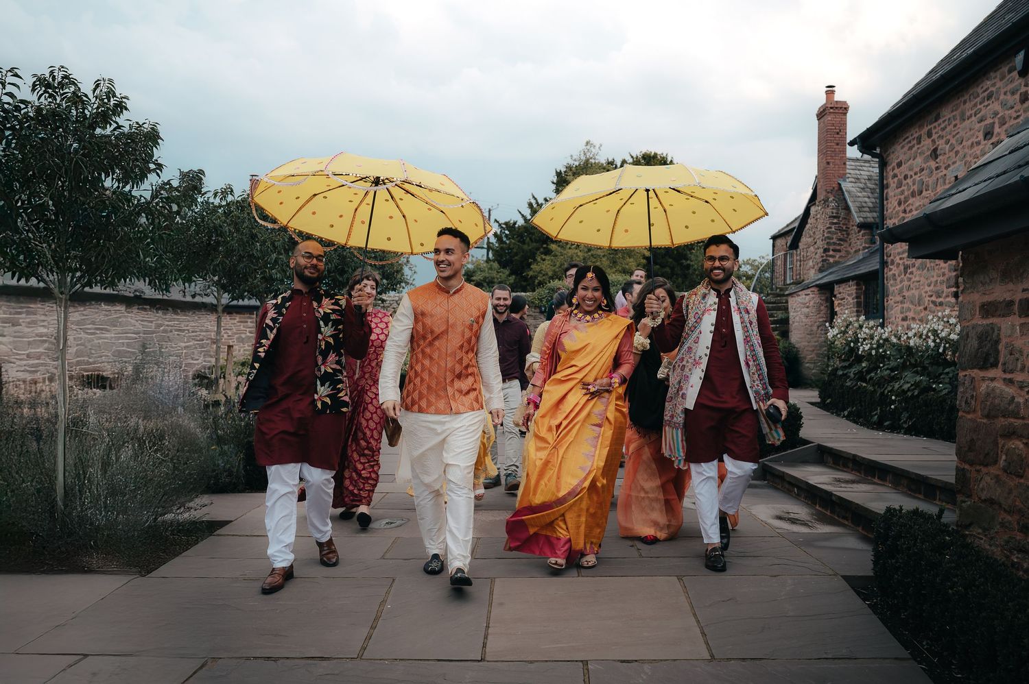 Buddhist monks in orange and burgundy robes walk along a path carrying yellow parasols during a procession.