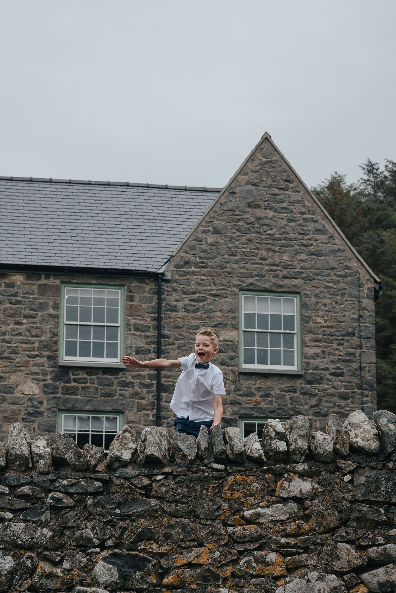 Child in white shirt standing on stone wall outside rustic building.