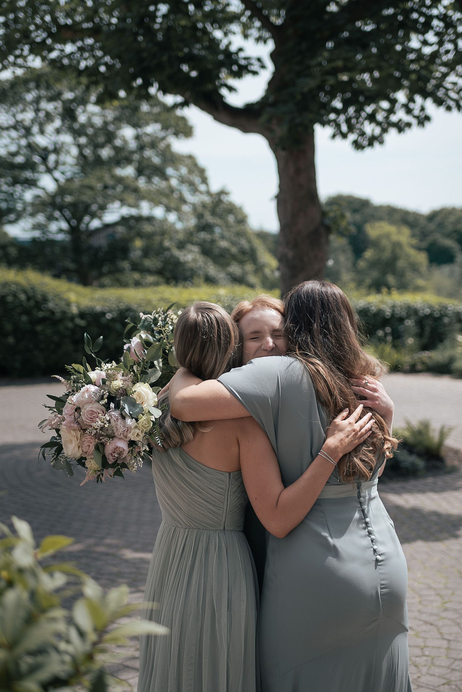 Three people share an emotional embrace while holding a floral bouquet outdoors.