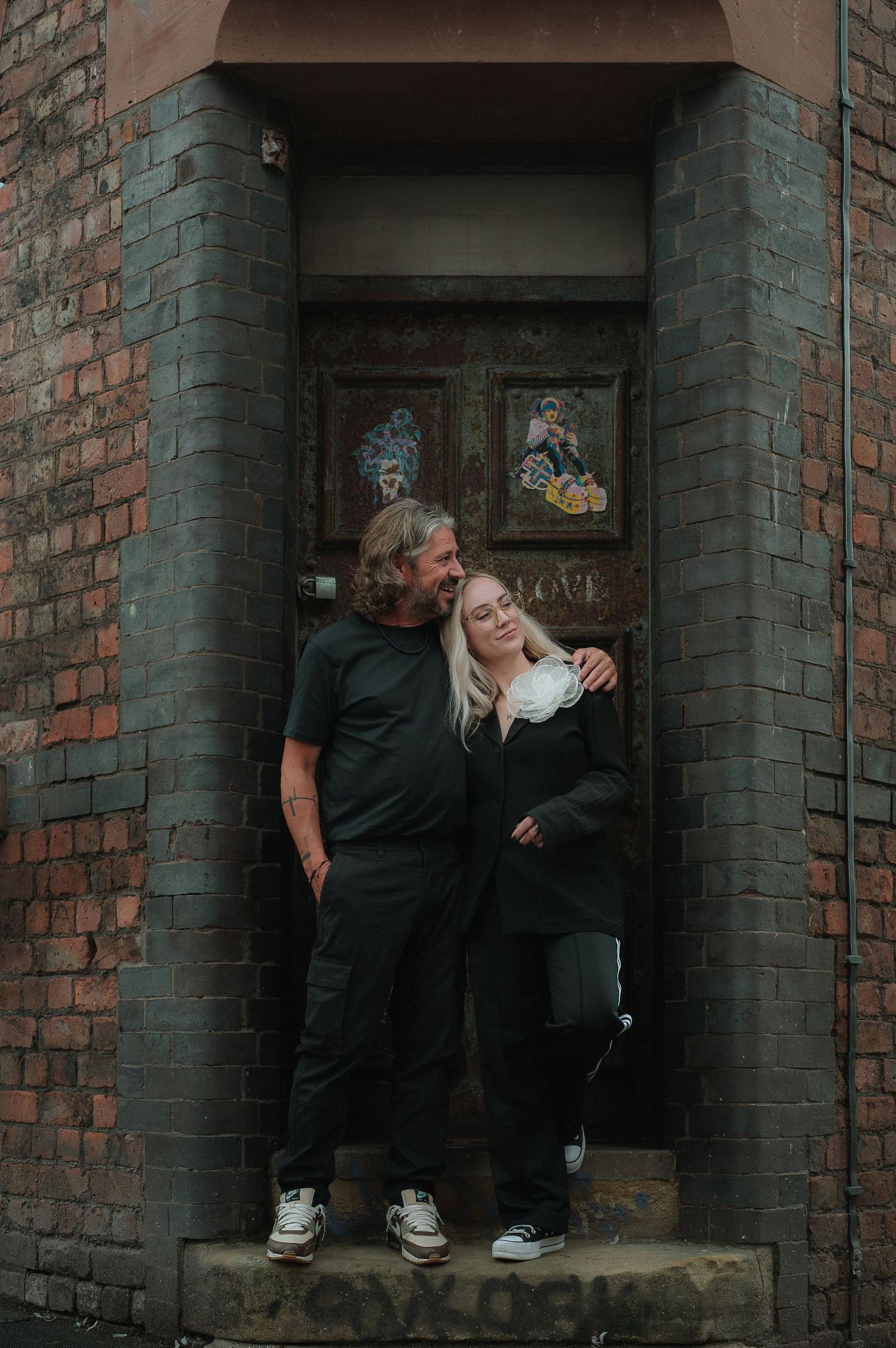 Two people share a tender moment in a brick doorway alcove with graffiti art in the background.