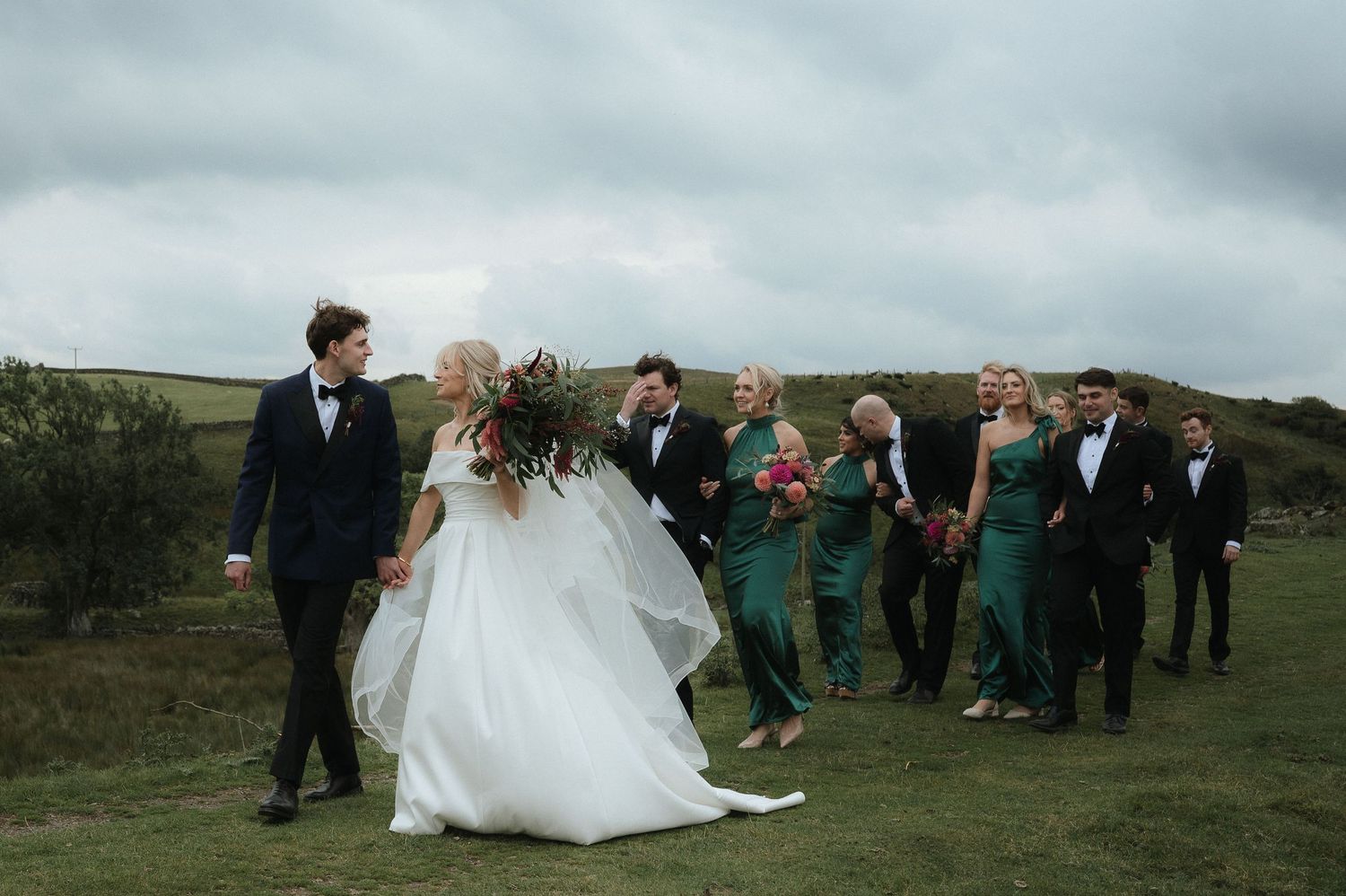 A wedding party gathers on a green hillside under cloudy skies during an outdoor celebration.