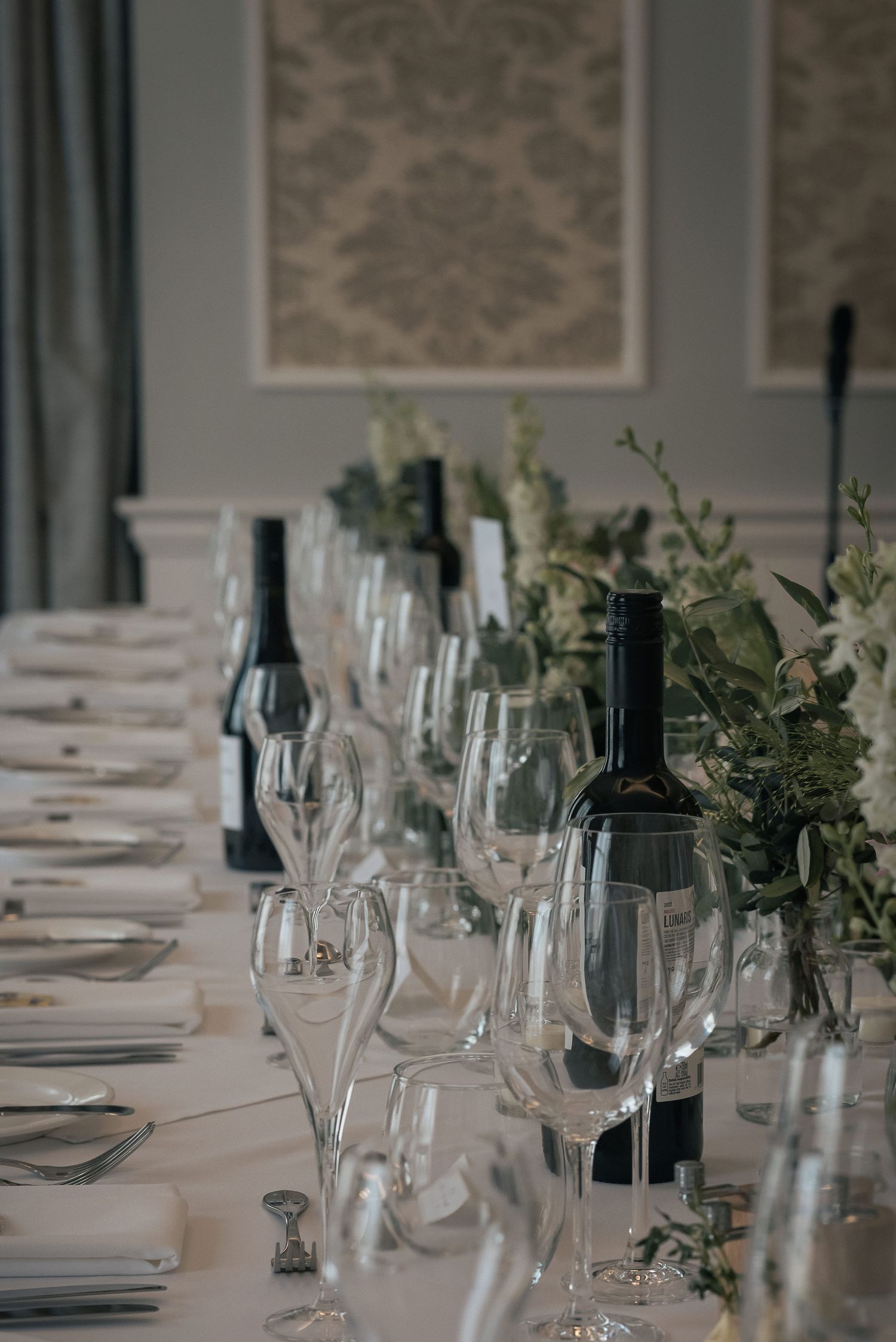 Elegant wedding table setting with wine bottles, glasses and greenery decorations along a white tablecloth.