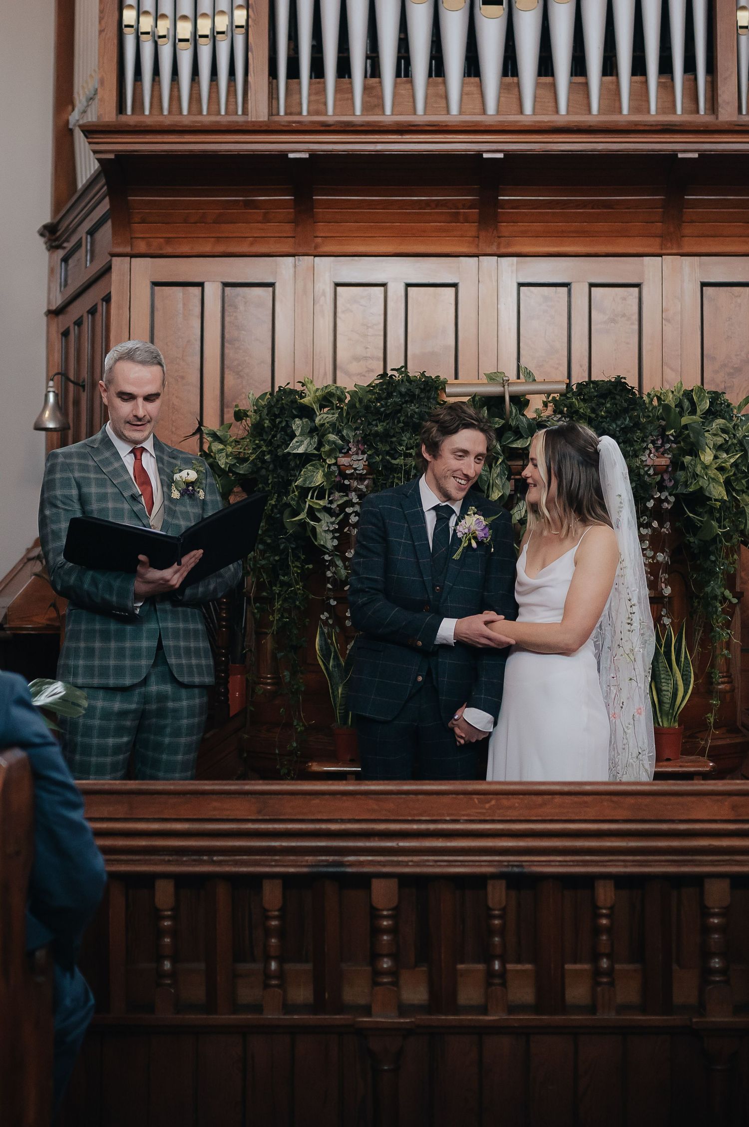 A wedding ceremony takes place in a wood-paneled room with an organ pipe backdrop.