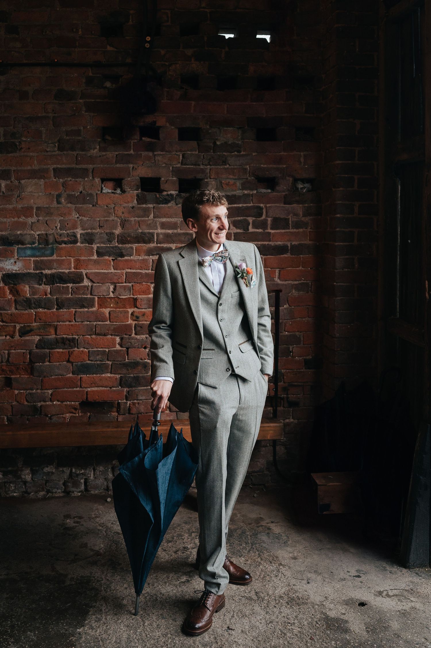 A well-dressed person in a light gray suit poses against a brick wall holding a blue umbrella.