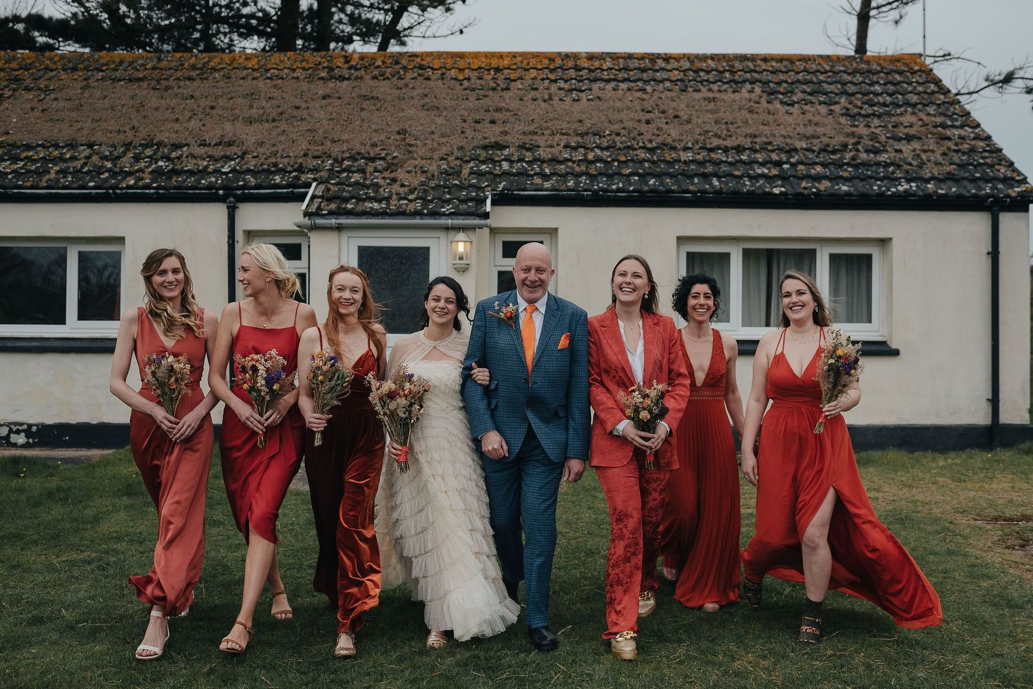 Wedding party posing outside rustic building in coral dresses and blue suit.
