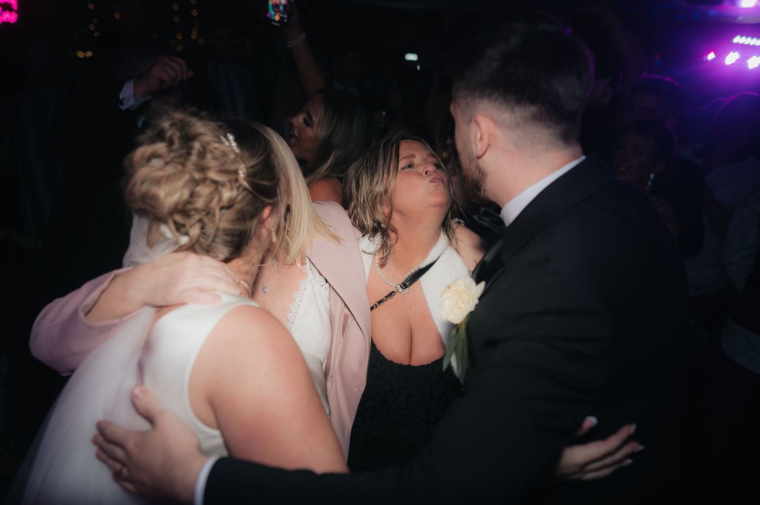 Wedding guests celebrate and dance together on a dark dance floor with purple lighting in the background.