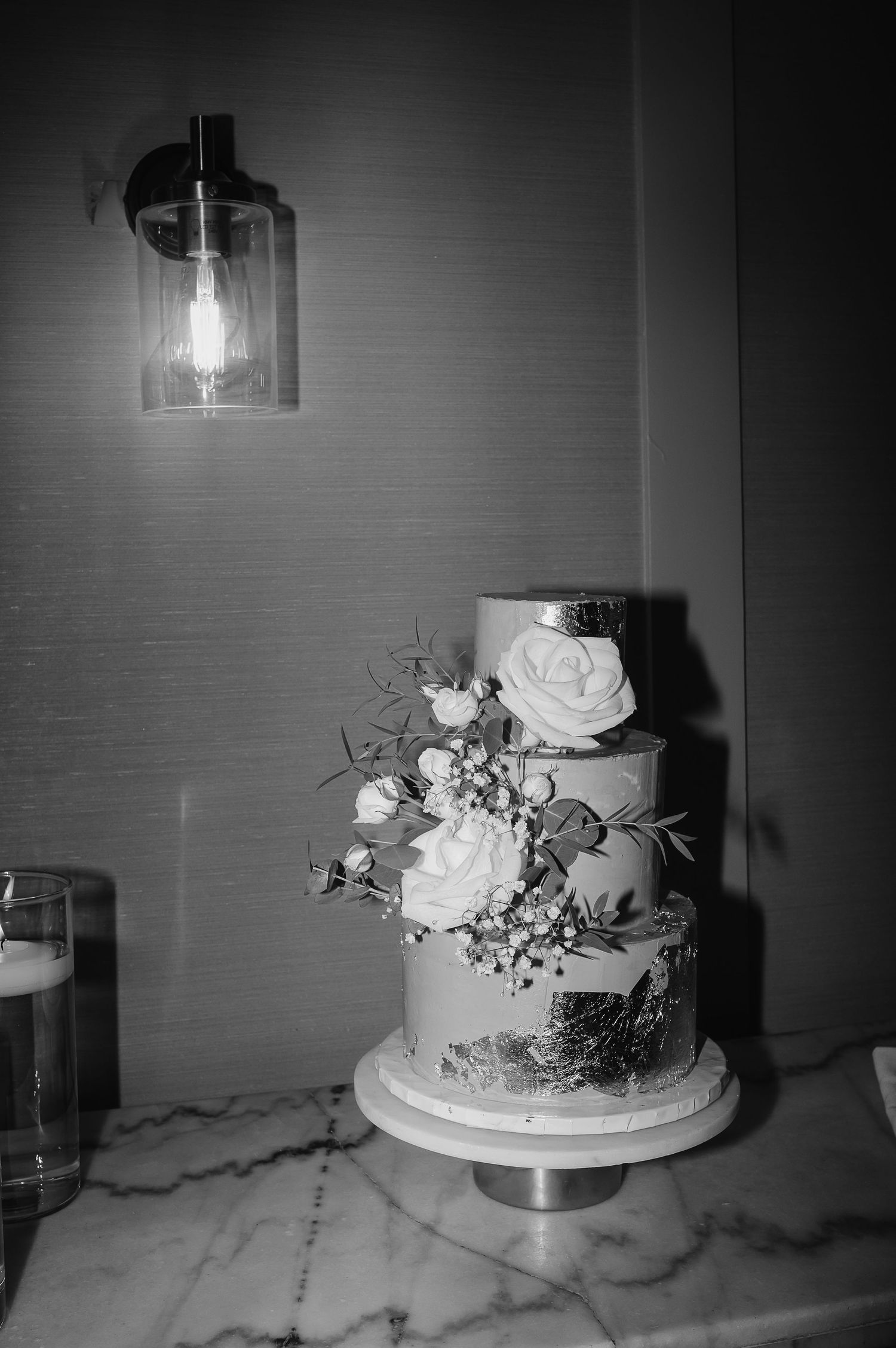 Elegant wedding cake with white roses and silver accents under pendant lighting.