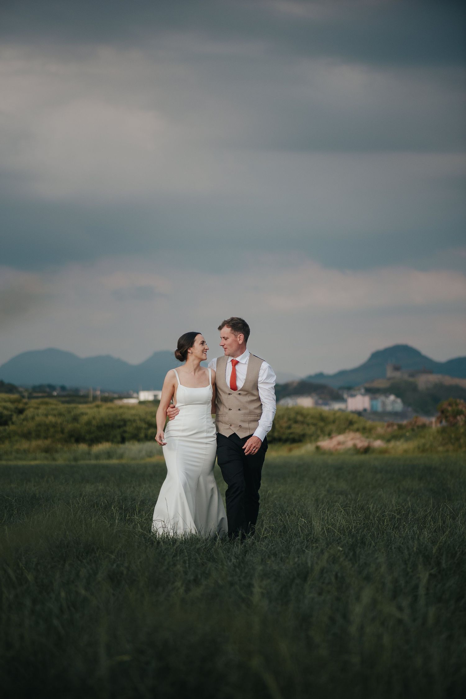 Newlywed couple stands in field with mountains and moody sky in background.