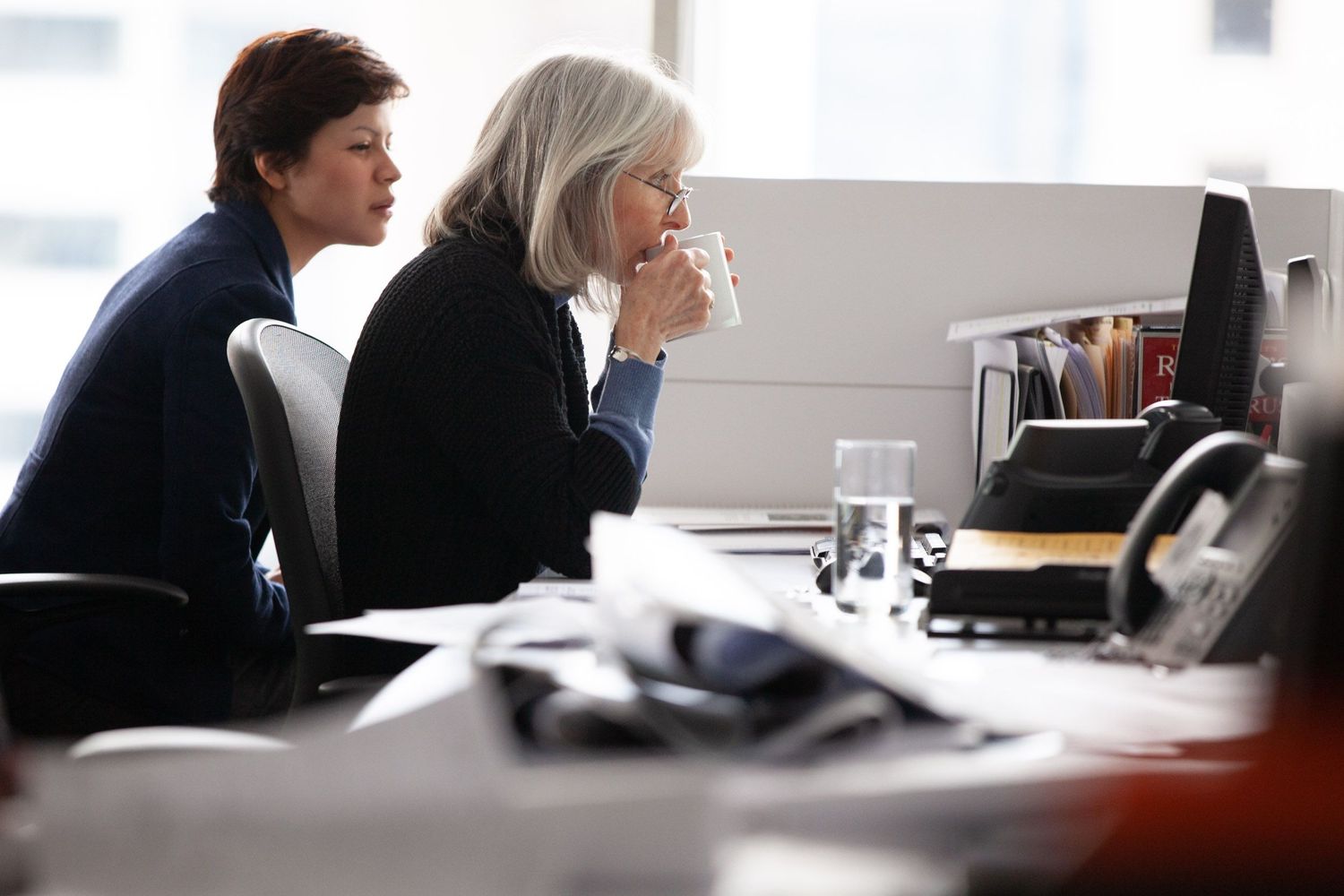 Two people sit at a desk in an office environment while one drinks from a mug during a business meeting.