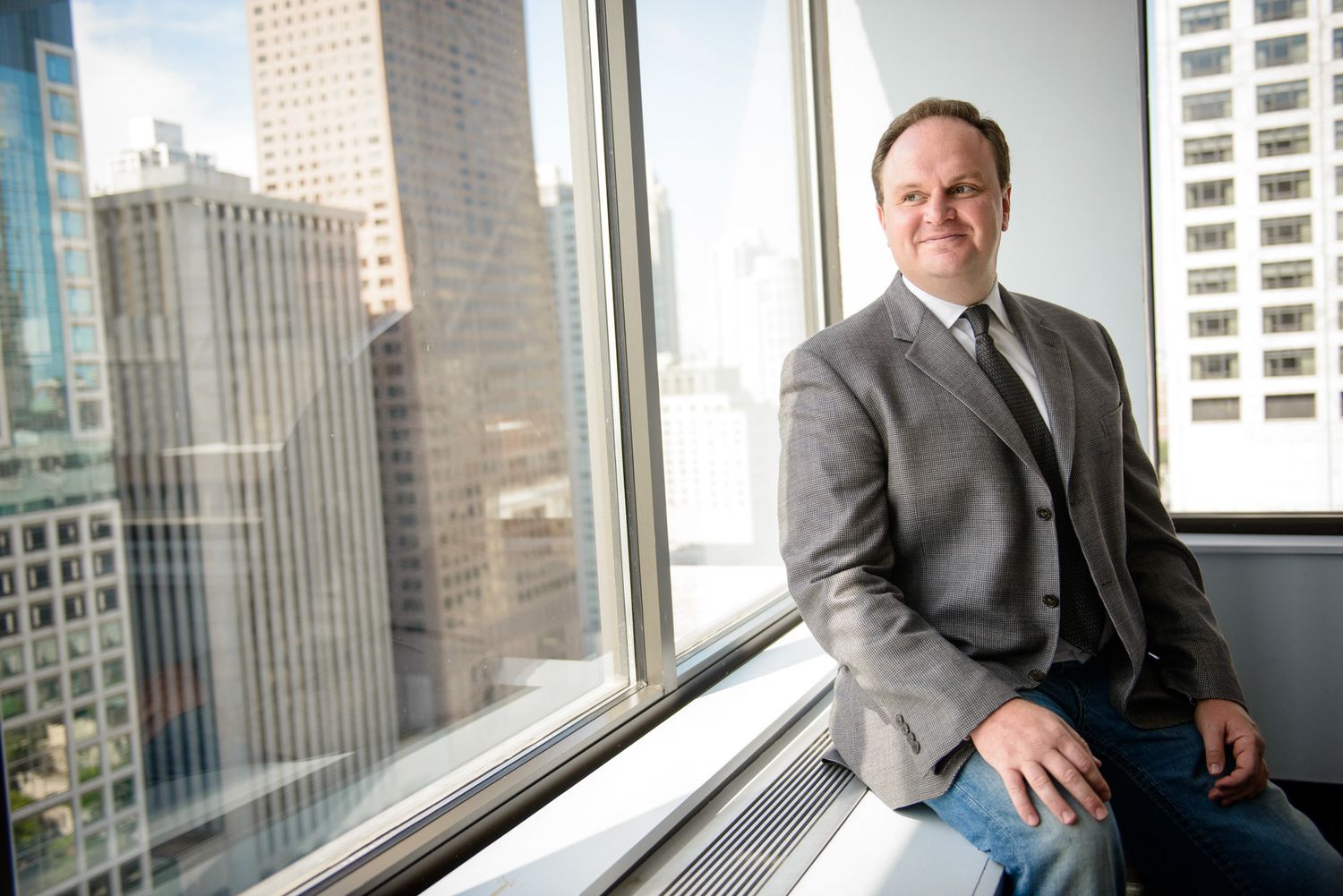 Business professional sits by large office windows overlooking urban skyscrapers in a modern corporate setting.