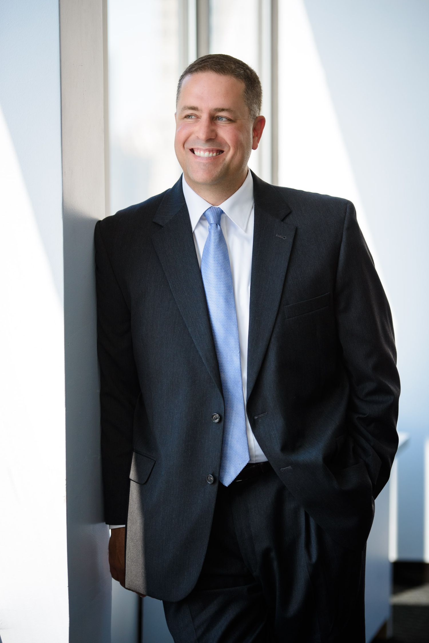 Professional business portrait of someone in a dark suit and blue tie standing against a light background.