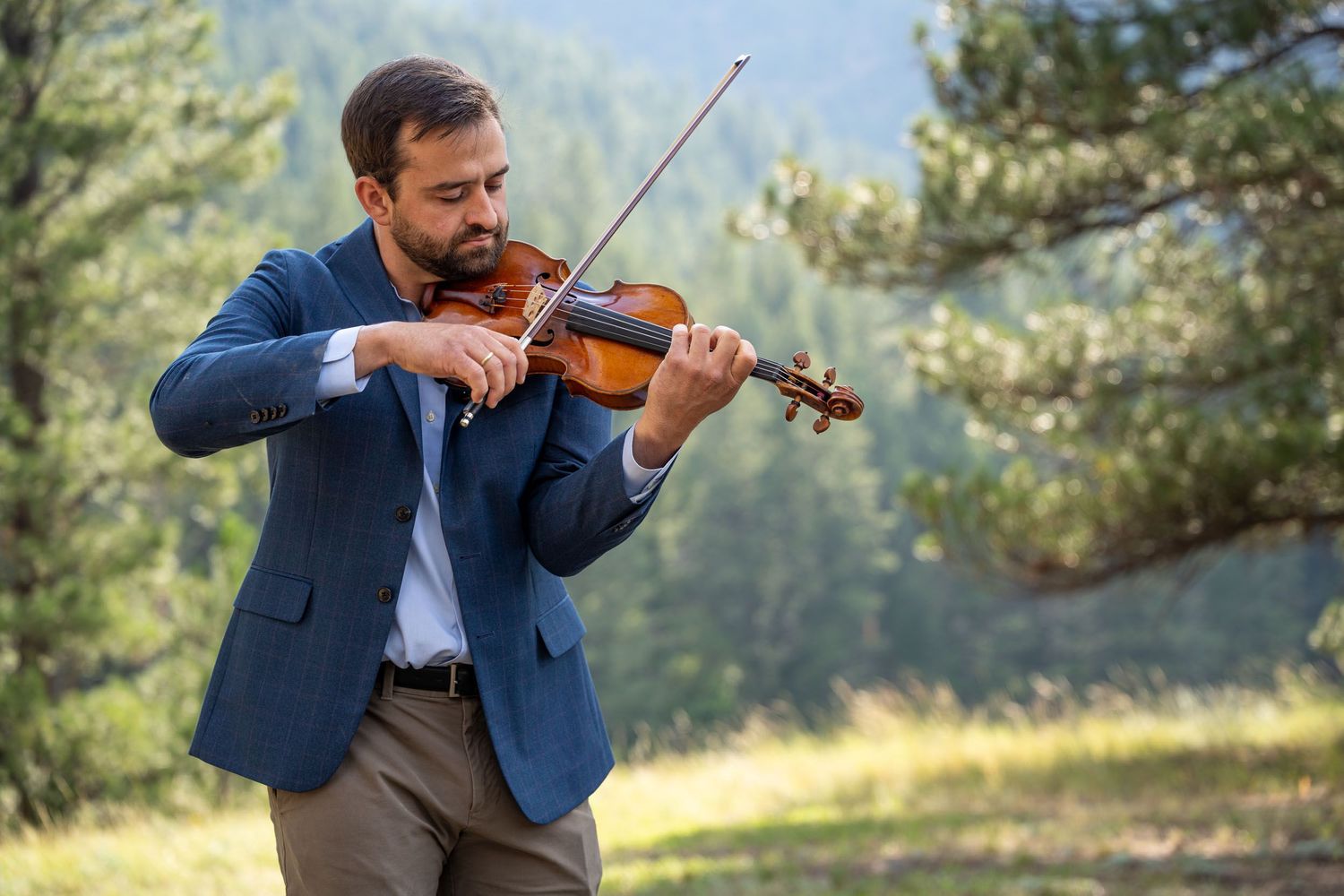 Violinist performing outdoors in a blue suit against a mountain landscape.