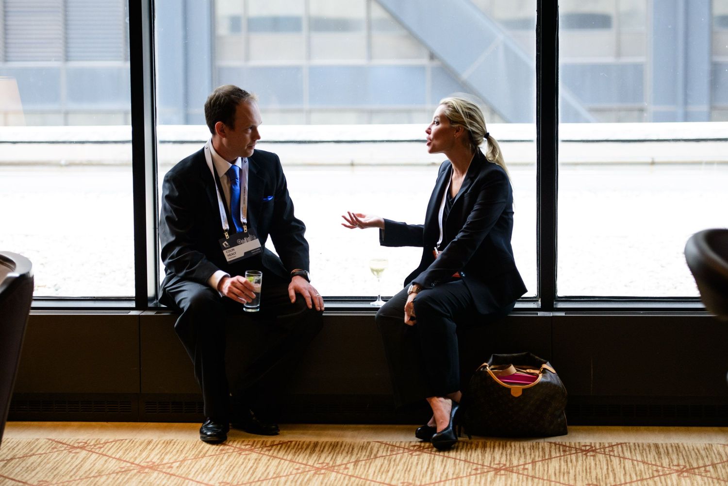 Business professionals in suits sit on the floor having an informal discussion in a modern office hallway with windows.