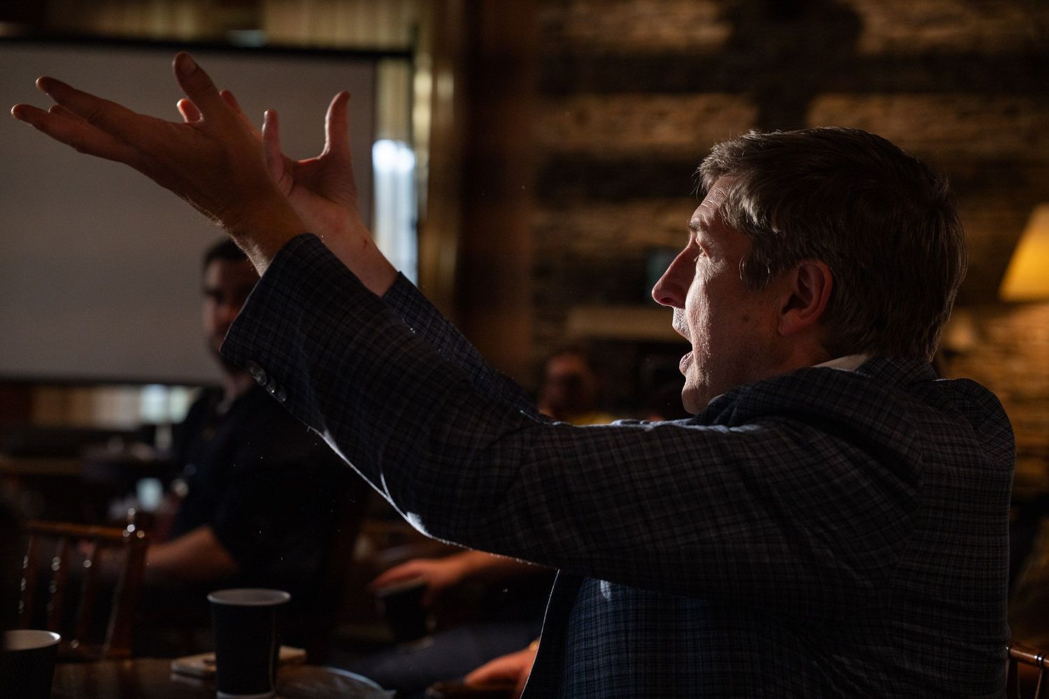 A person in a dark sweater gestures with raised arms while sitting in a dimly lit rustic interior space.