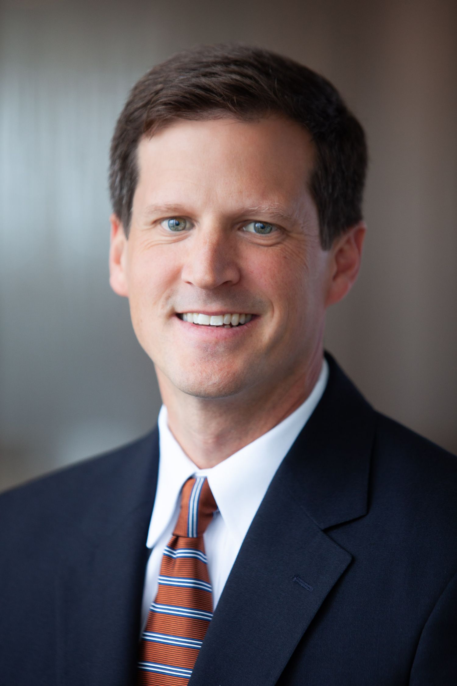 Professional headshot of someone wearing a navy suit and orange striped tie against a blurred background.