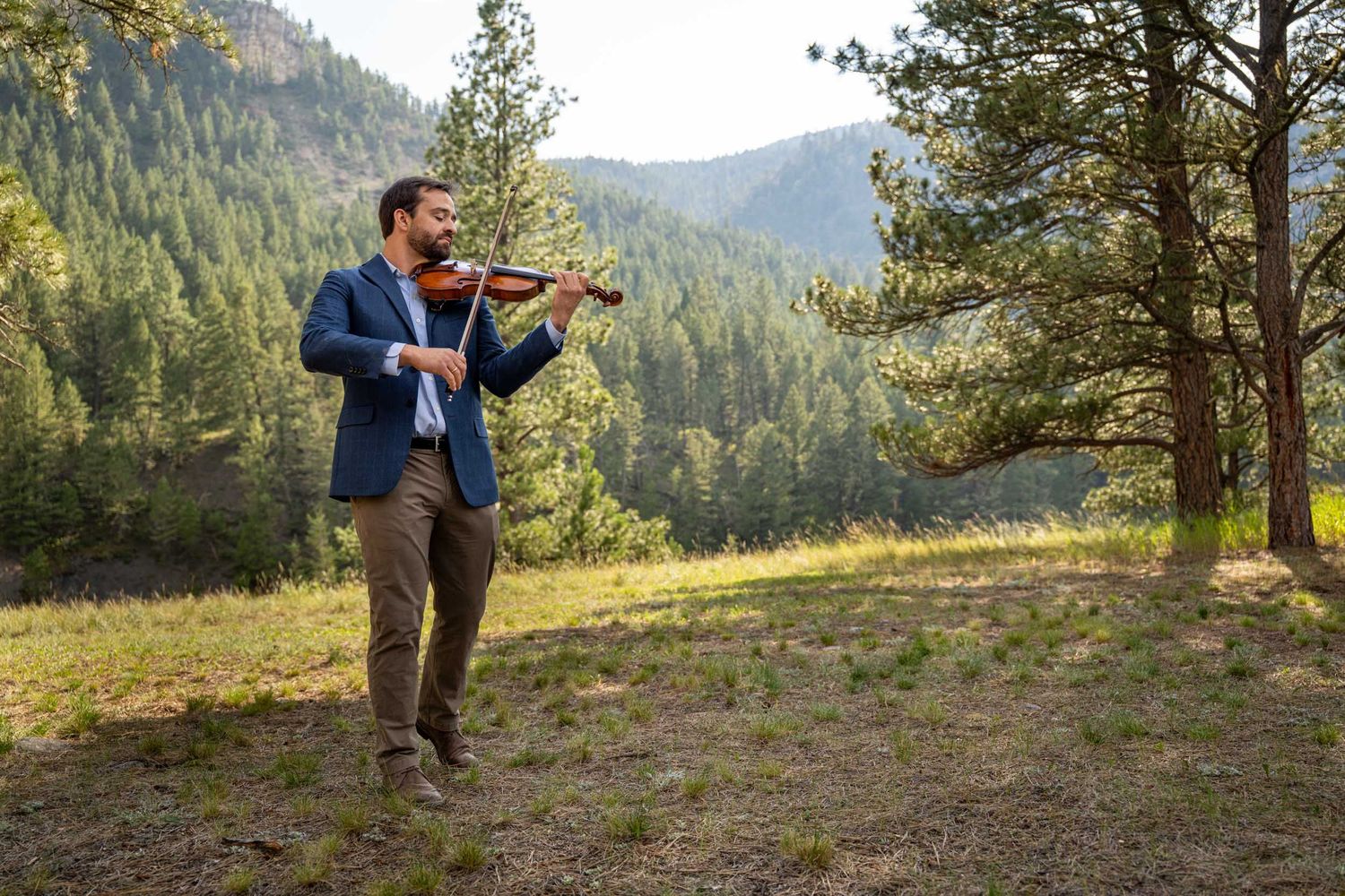 A violinist performs in a scenic mountain forest setting during golden hour light.