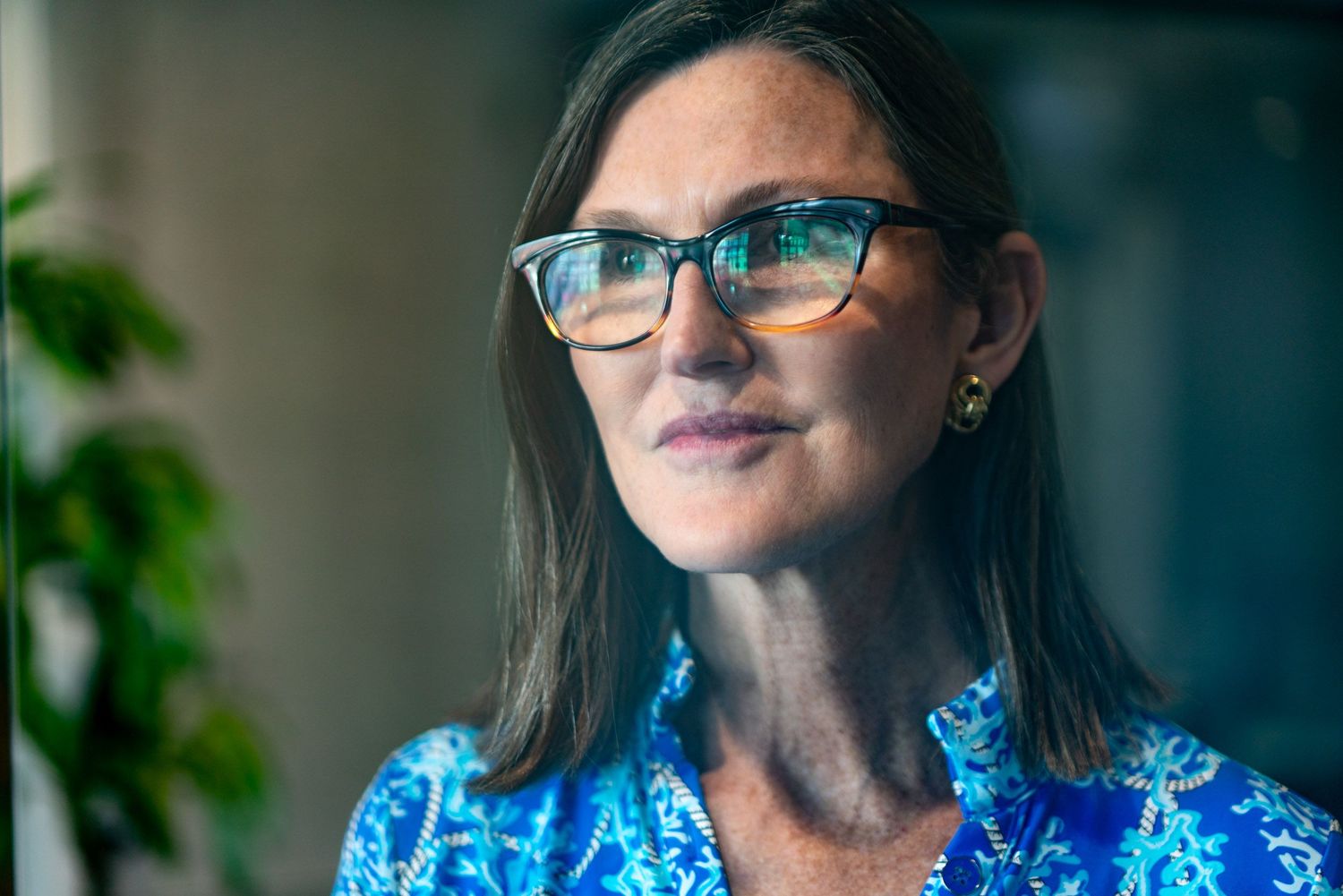 Person in blue floral dress and glasses smiling while sitting indoors near a window.