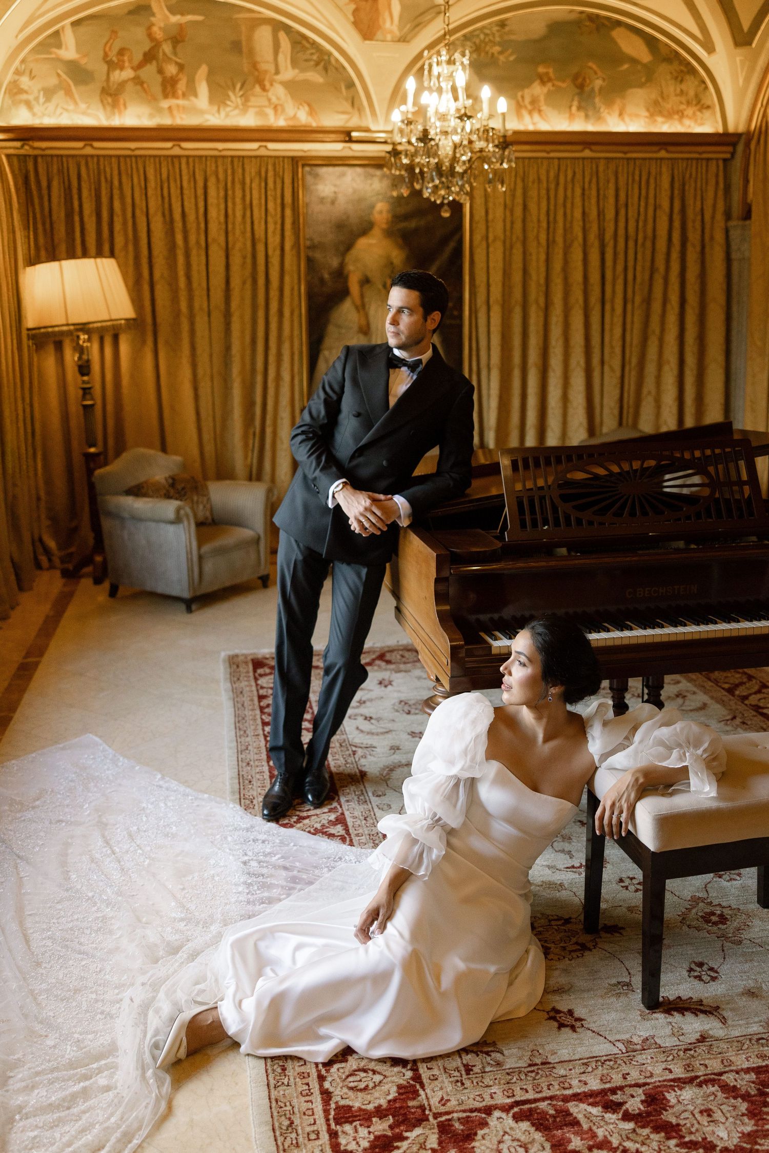 Elegant bride in white gown sits on floor beside groom in black tuxedo near grand piano in ornate room.