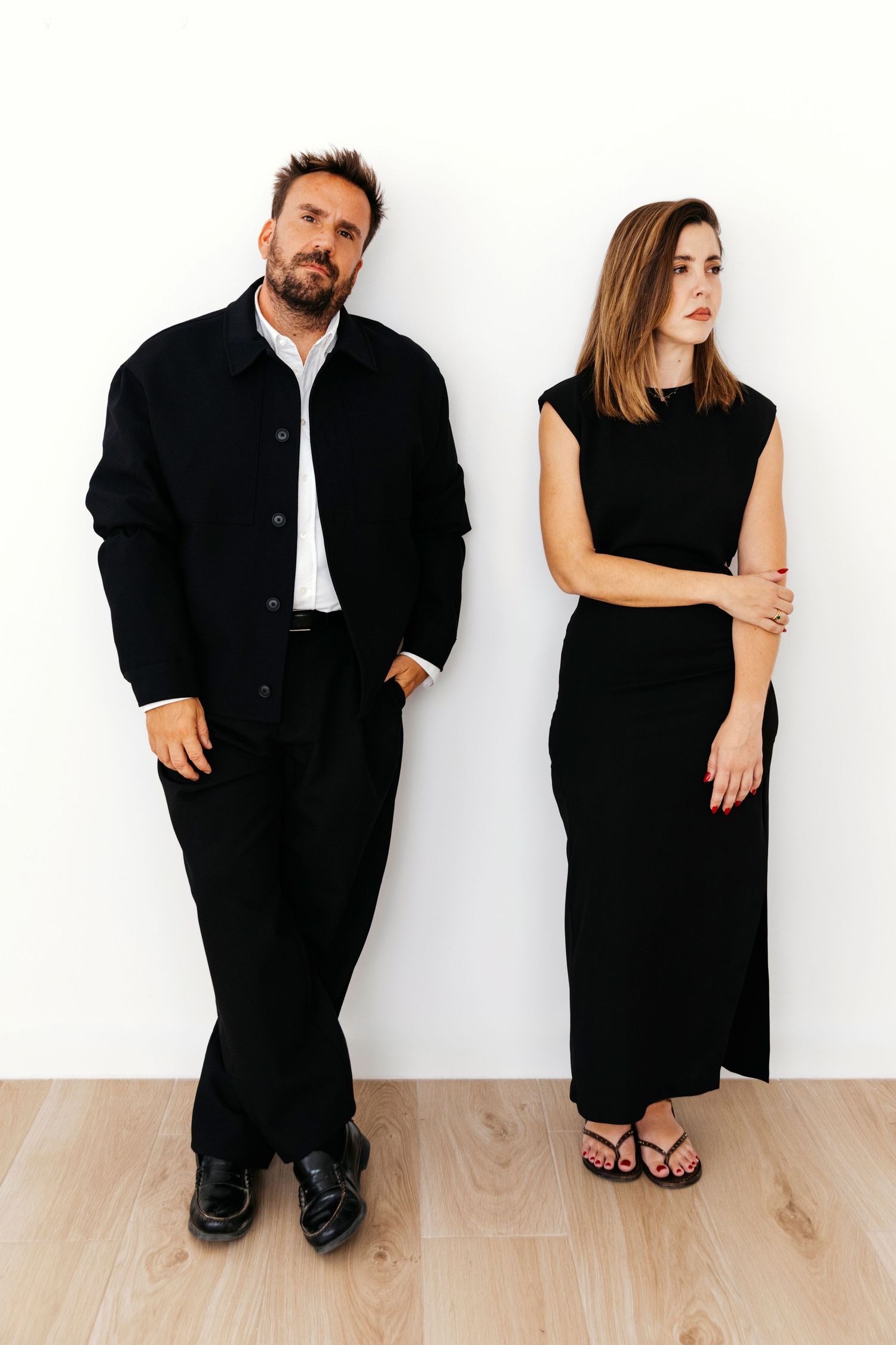 Artistic black and white portrait series showing two people in black attire sitting and posing against a white wall.