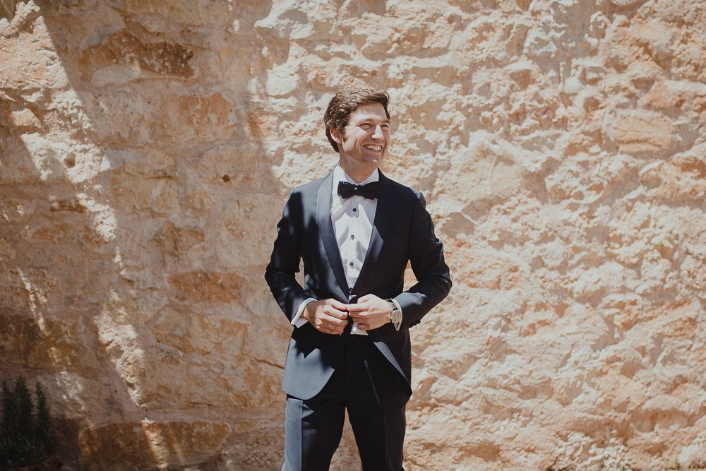 Figure in black tuxedo and bowtie poses against stone wall in sunlight.