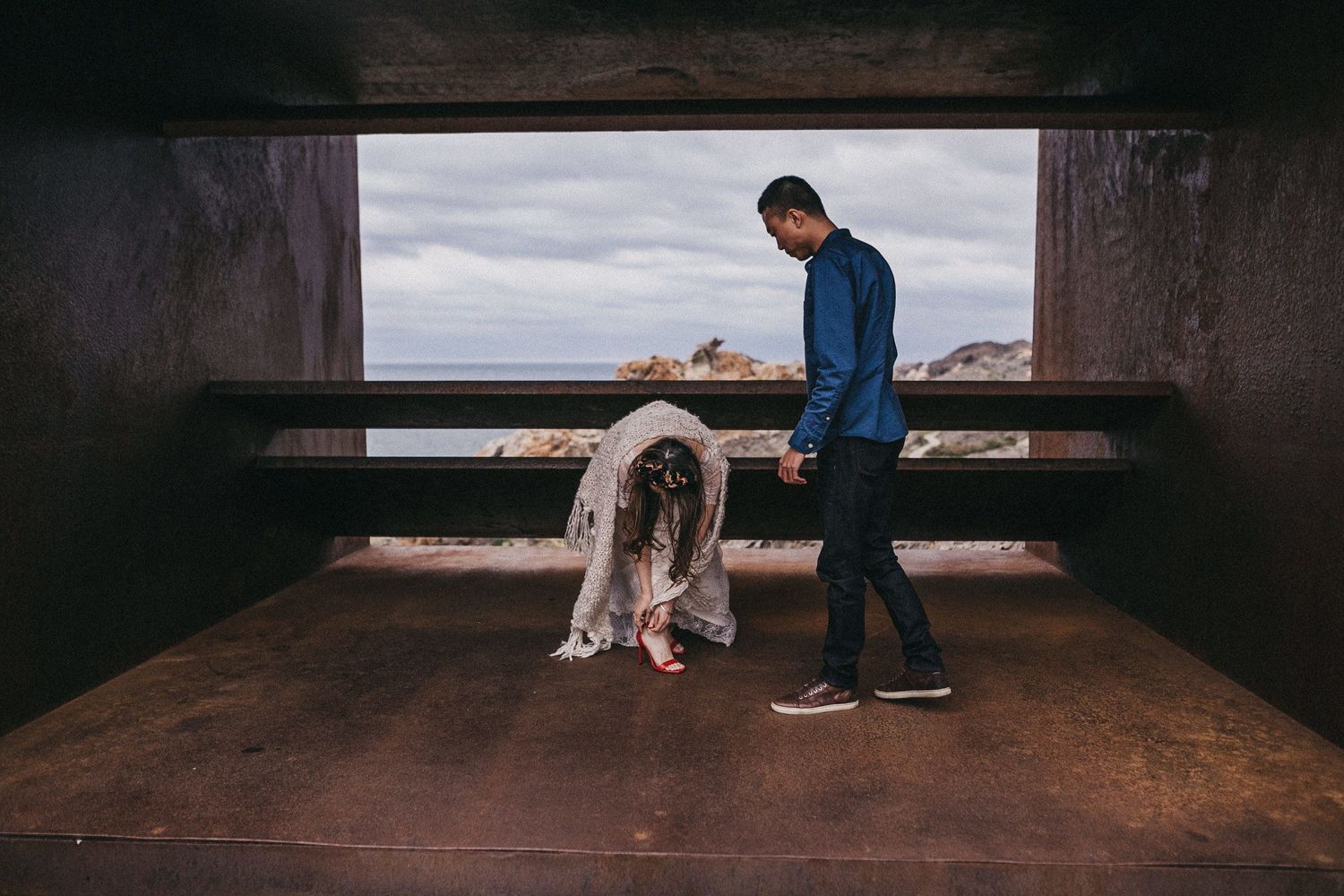 Two people share a moment in a modern viewing platform against a cloudy coastal backdrop.