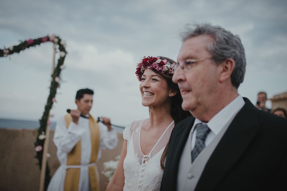 Wedding ceremony under floral arch with Mediterranean landscape in background.