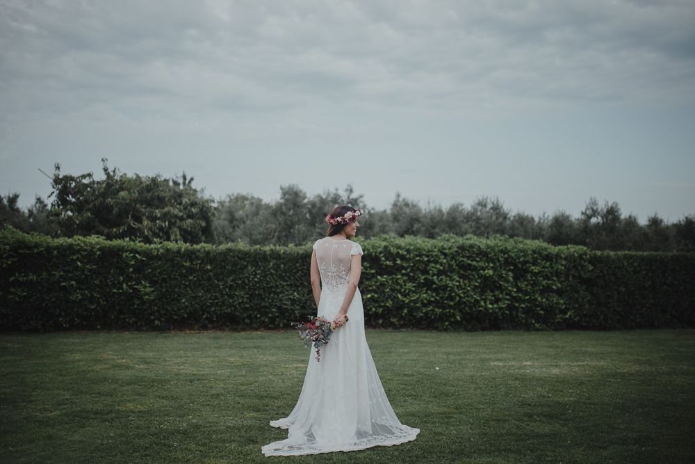 A bride in a white dress stands in front of manicured hedges holding bouquet behind back.