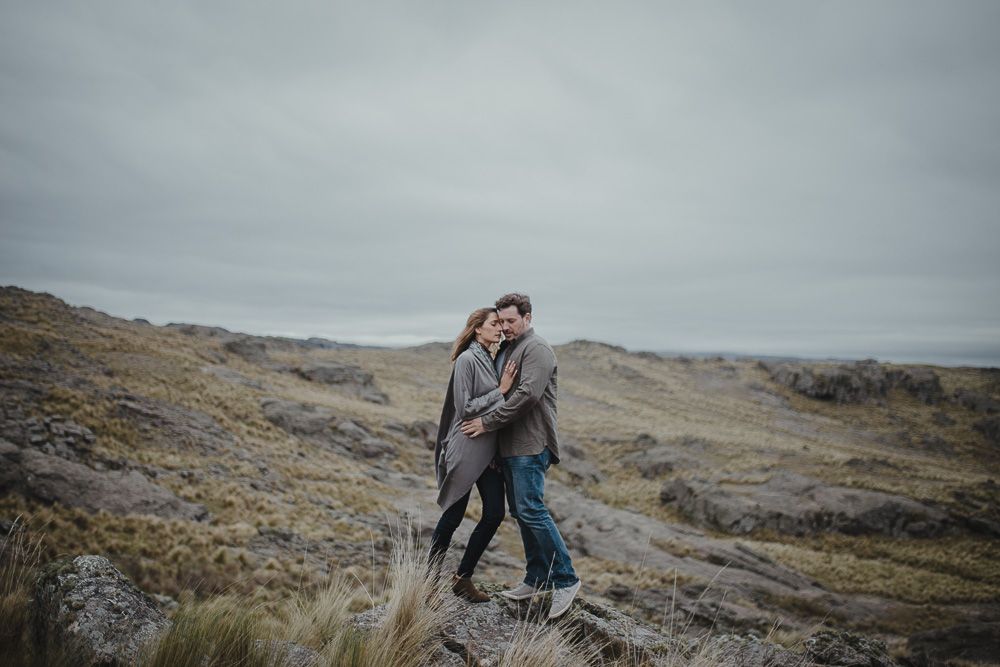A couple shares a tender moment while standing among tall grasses on a cloudy hillside.