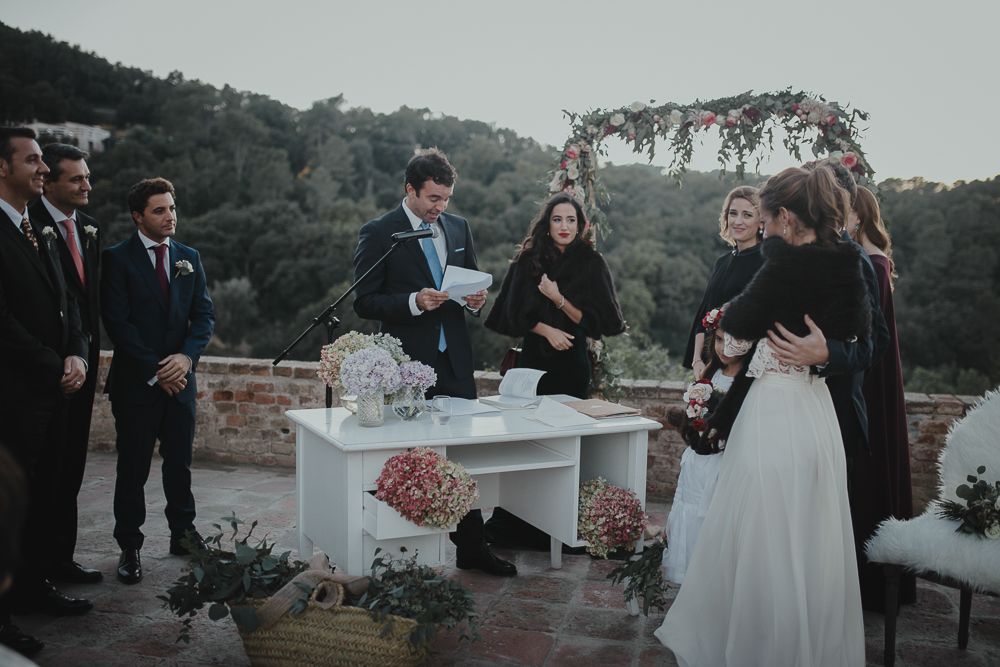 A wedding ceremony takes place outdoors at dusk with attendees gathered around a white table for document signing.