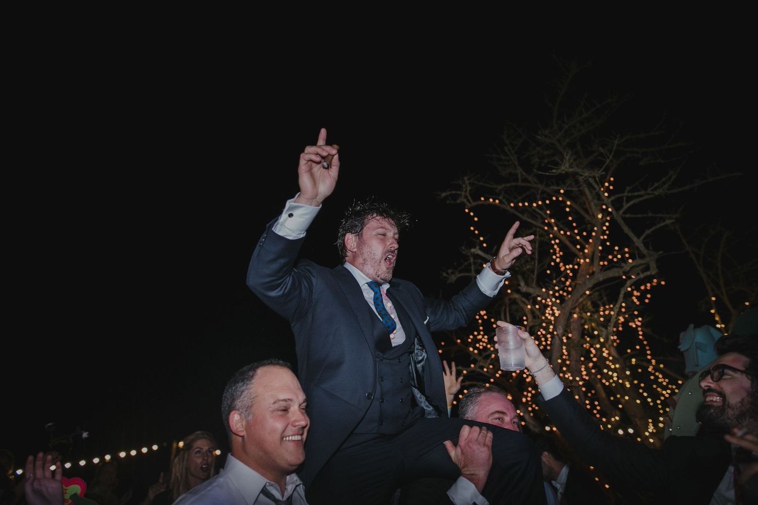 Wedding guest being lifted up on shoulders during nighttime outdoor celebration.