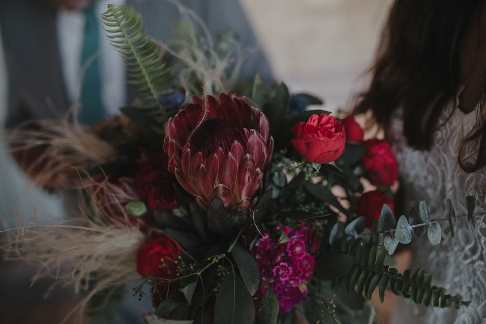 A moody floral arrangement with dark red protea flowers and greenery in dramatic lighting.