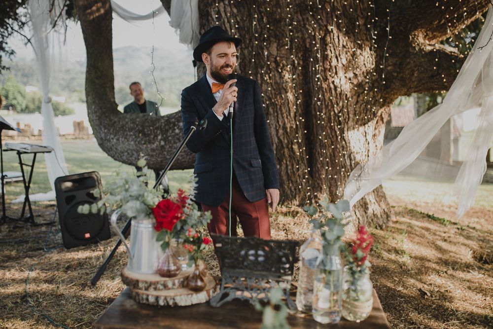 A wedding speech being given next to a decorated dessert table under a large tree.