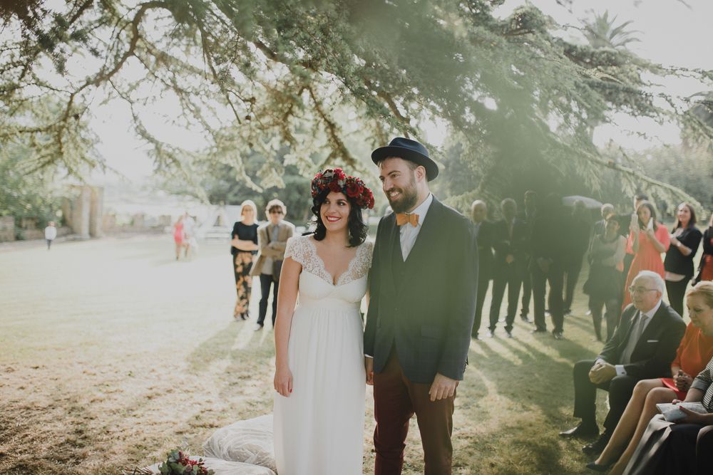 A couple stands together during their outdoor wedding ceremony under tall trees.